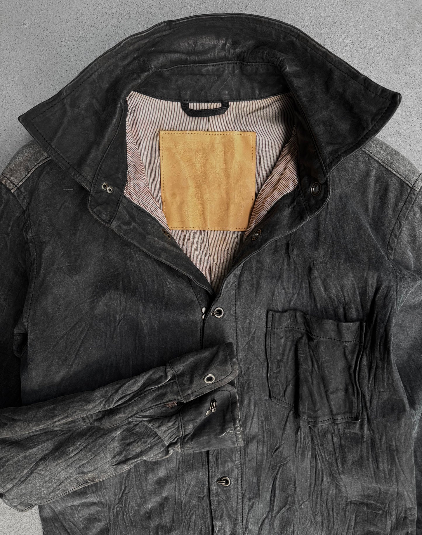 A black, washable wrinkle nubuck leather shirt by Kyoji Maruyama from the early 00s is laid flat with a high collar, front pocket, striped lining, tan inner patch, and the left sleeve folded across the body.