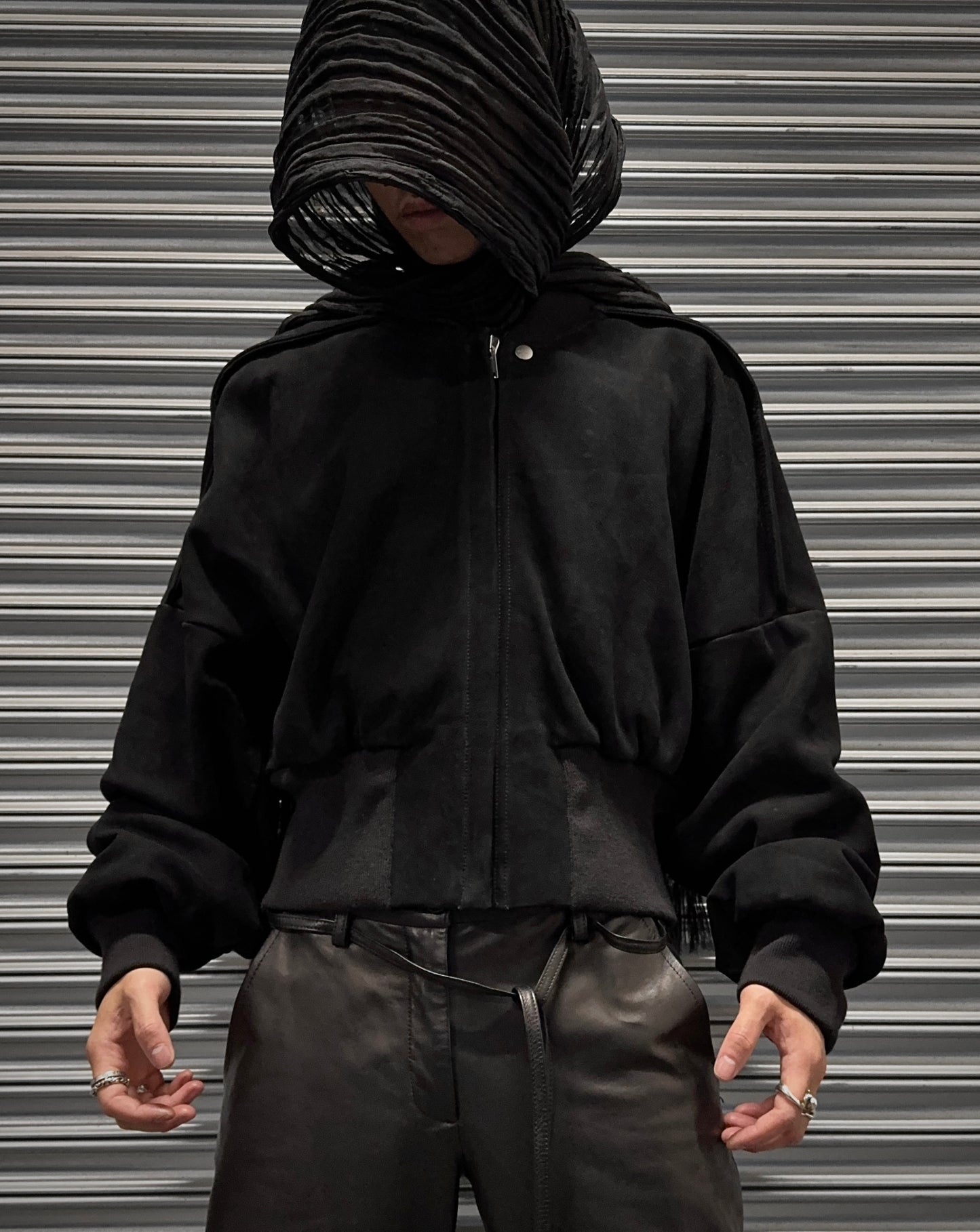 A person stands before a metal shutter in the Rick Owens SS24 “LIDO” Nubuck Leather Cropped Flight Bomber Jacket, loose black pants, and a textured black head covering, their ring-adorned hands finishing the avant-garde look.