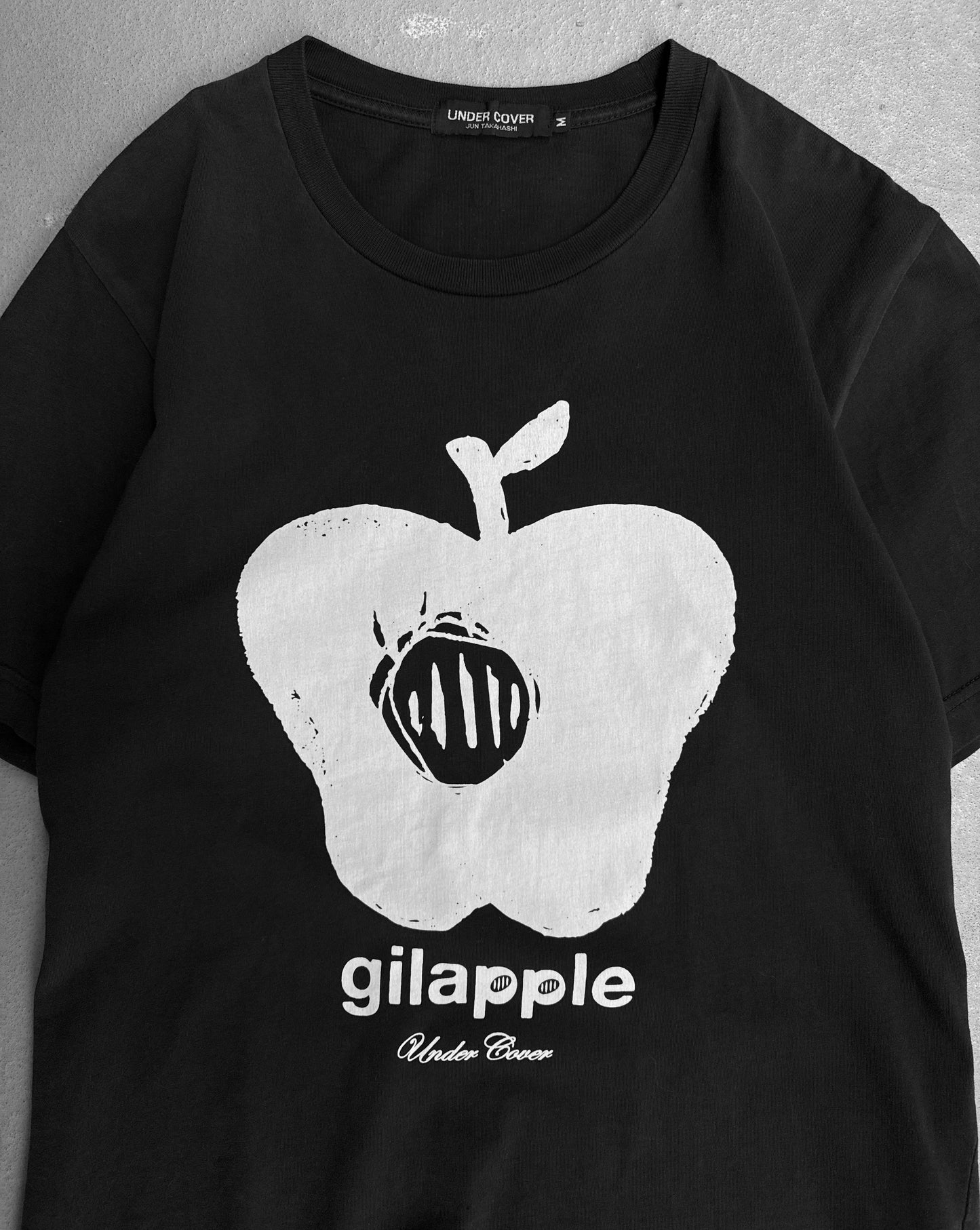 The Undercover SS16 ‘Gilapple’ Graphic Printed T-shirt is black with a bold white apple graphic and black lines, featuring “gilapole” and “UNDERCOVER” text beneath, reflecting Jun Takahashi’s signature streetwear style.