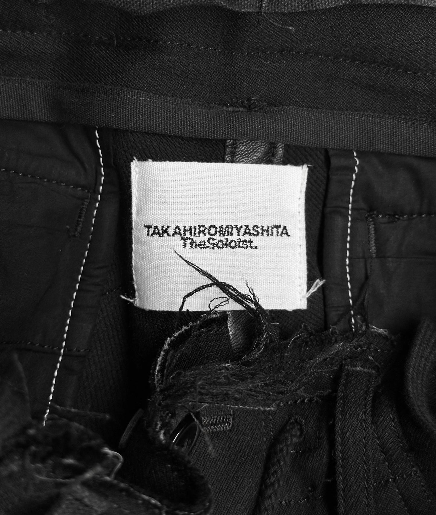 Close-up of a clothing label on black pants, featuring TakahiroMiyashita TheSoloist. in bold text against white. These avant-garde AW15 Calf-trimmed Wool Bondage Straps Cargo Pants have black stitching and artfully frayed edges near the label.