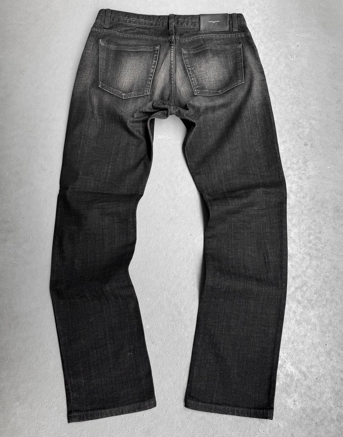The Surface To Air SS14 Faded Black Bootcut Skinny Denim is displayed flat on a grey surface, showcasing the back with visible pockets and subtle seam fading—ideal for those who value urban style in modern menswear.