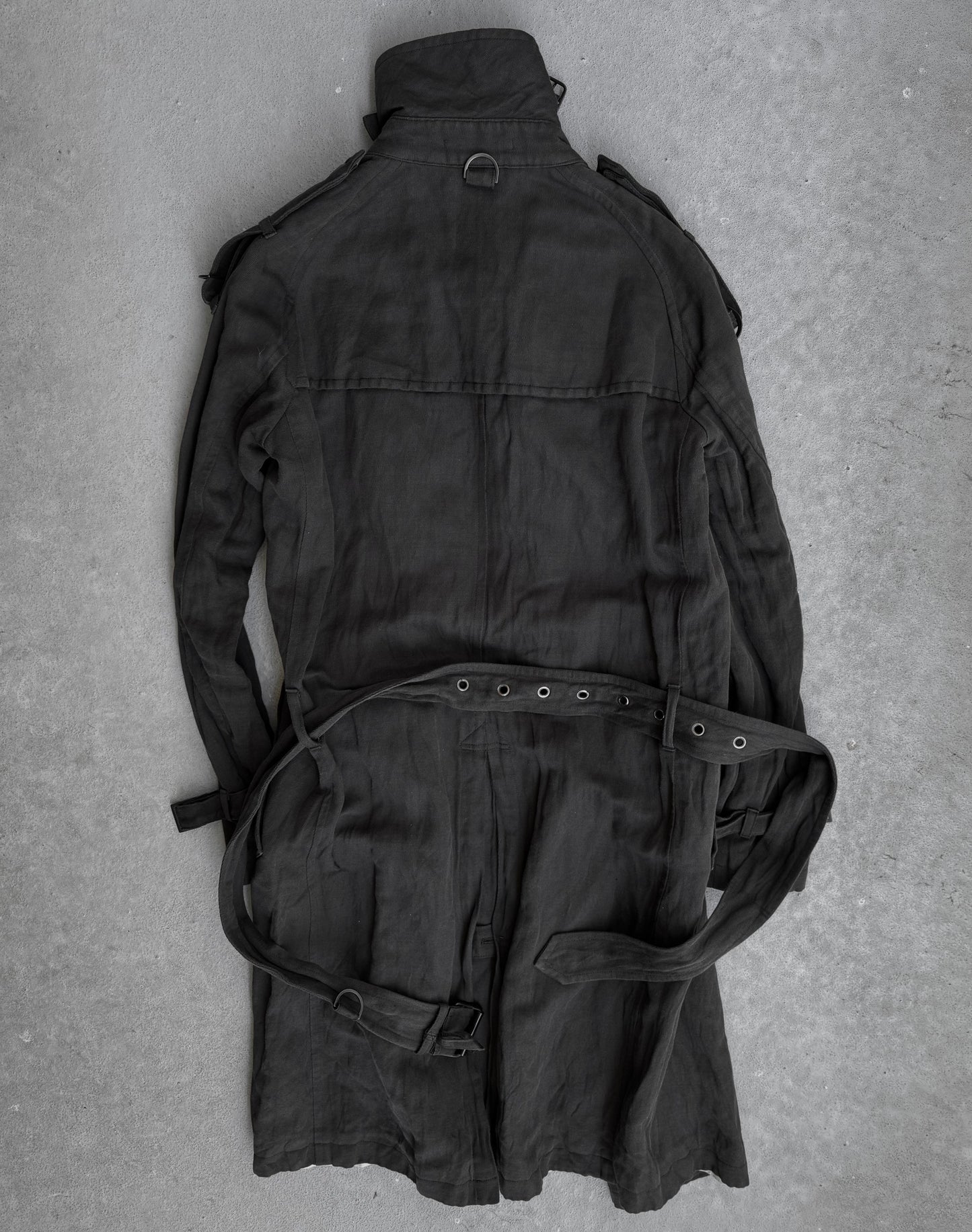 The ATTACHMENT by Kazuyuki Kumagai Raglan Doctor Trench Coat is laid flat on a gray concrete surface, displaying its back and sleeves—a sleek piece from Attachment by Kazuyuki Kumagai.