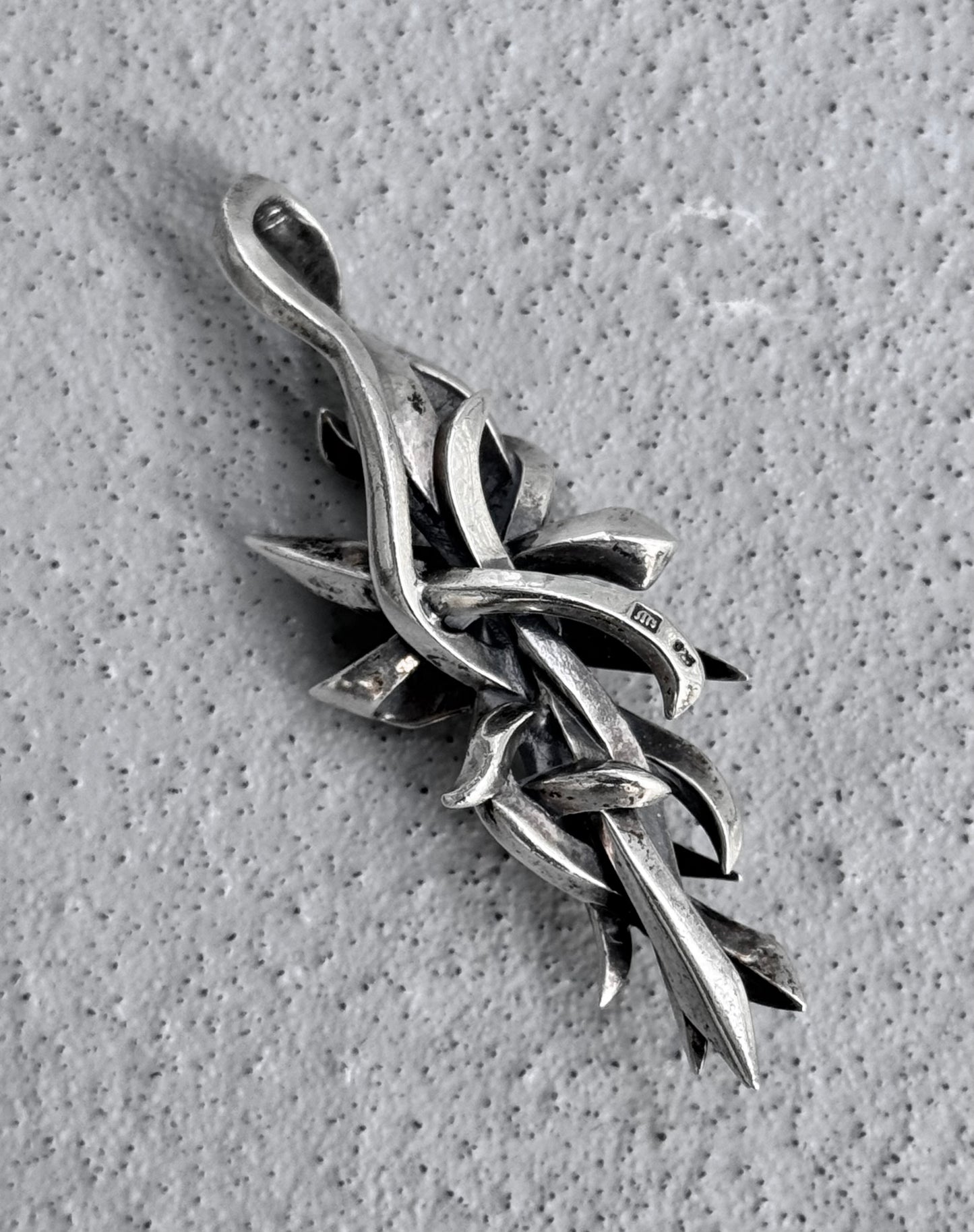 Close-up of M’s COLLECTION (BLESS) Early 00s Silver Arabesque Cross Pendant by BLESS, featuring intertwining vine-like designs on a textured gray background. Its dynamic and elegant curved, pointed elements reflect the spirit of sustainability.