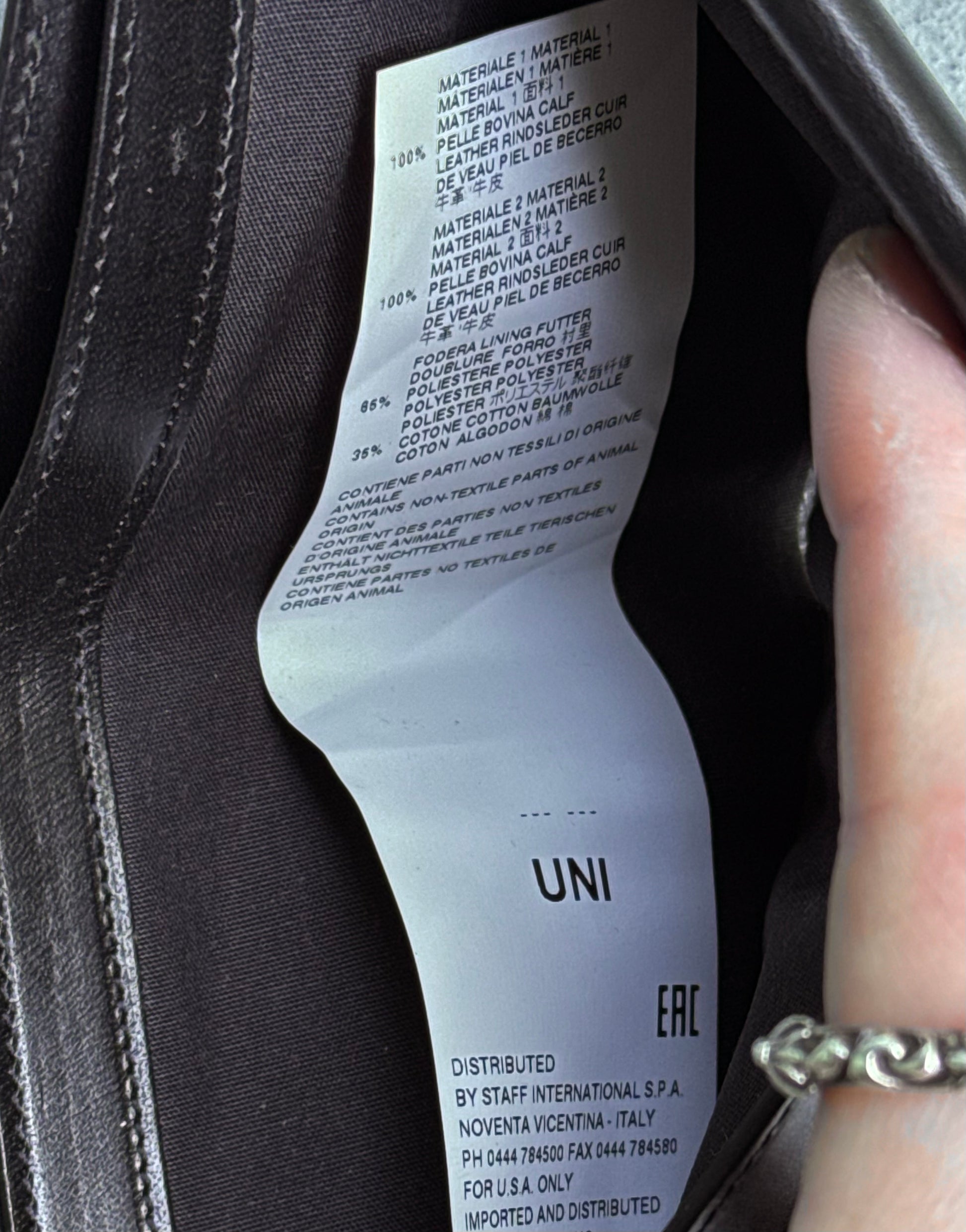 A hand with a silver ring holds the label inside a Maison Martin Margiela AW15 Distressed Calfskin Leather Bi-fold Wallet, showing material composition and care instructions. Condition 8/10, gently used.