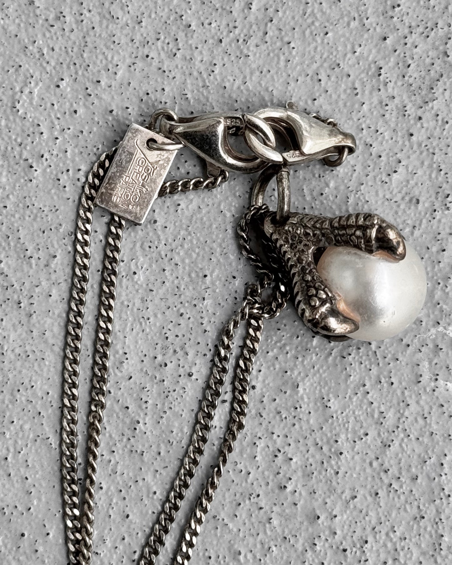 The YOSHiKOCREATiON Early 00s Silver Eagle Claws Pearl Chain by Yoshiko Creations showcases a claw gripping a pearl-like orb. Its interlocking chain links highlight innovative design, with a rectangular tag near the clasp, all resting on a speckled gray surface.