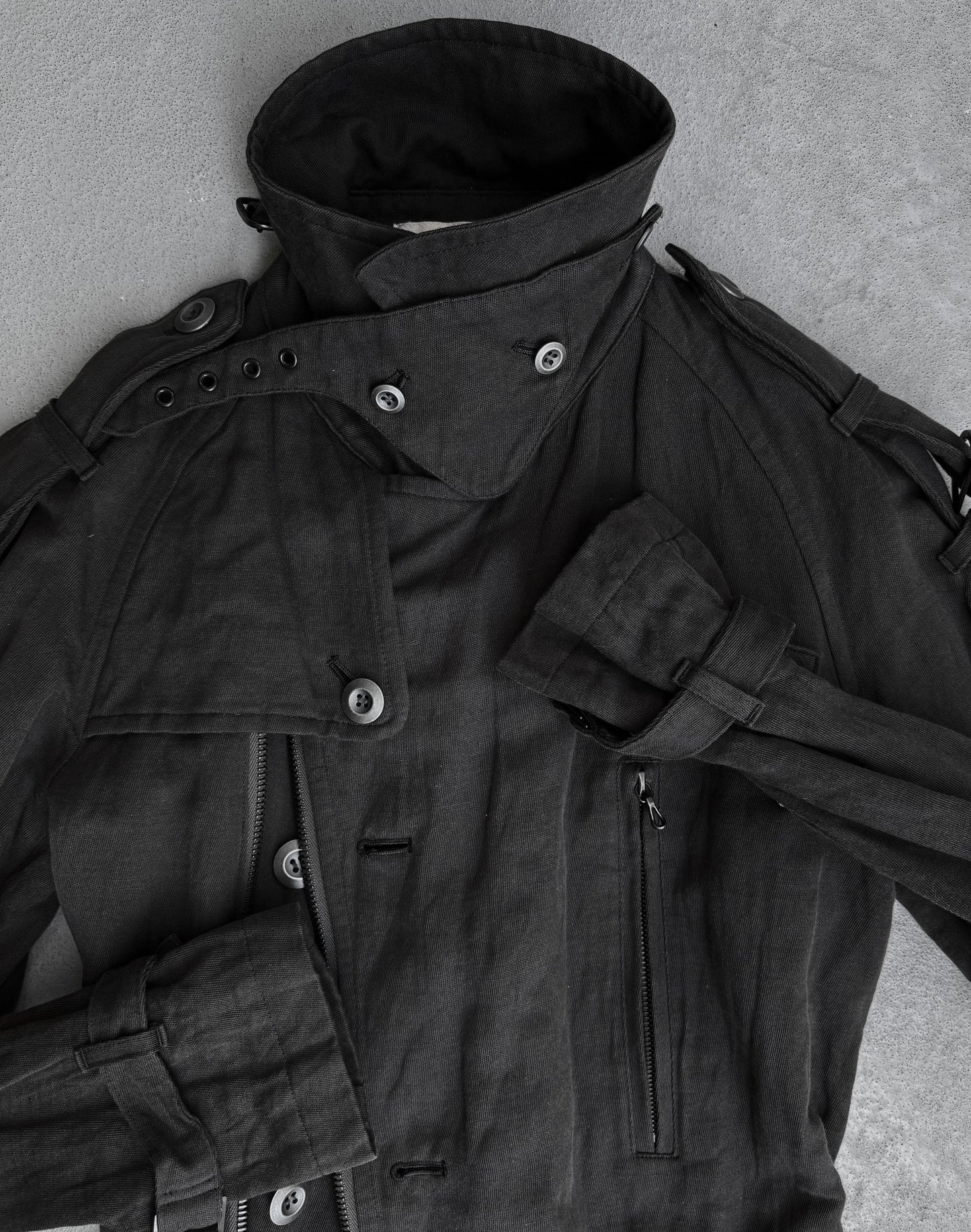 A dark grey ATTACHMENT by Kazuyuki Kumagai Raglan Doctor Trench Coat with a high collar, multiple buttons, epaulets, strap details, and a zippered pocket lies flat on a light grey surface—a refined menswear piece.