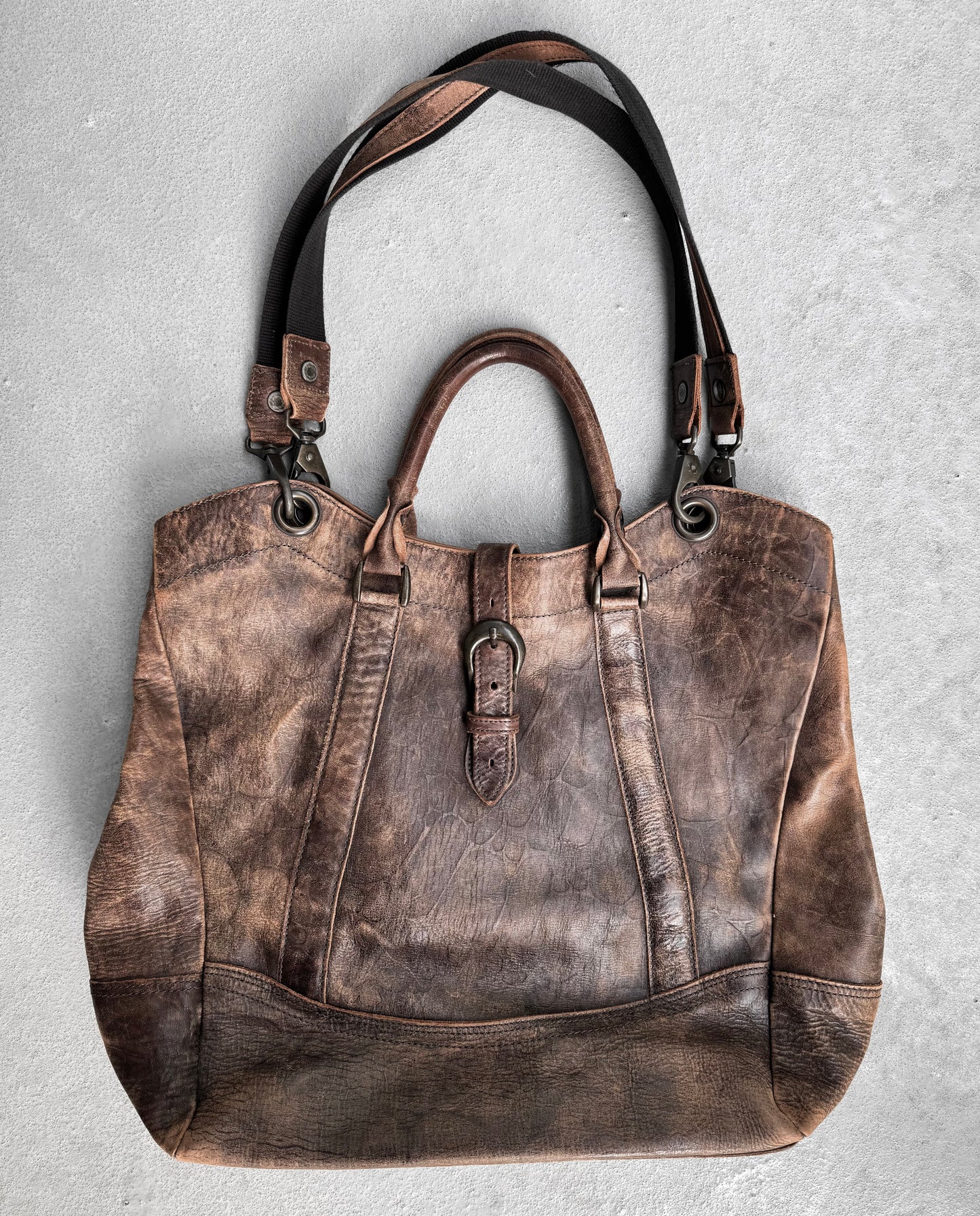 A vintage alfredoBANNISTER Early 00s Faded Brown Cowhide Tote Bag, crafted from Japanese leather with a weathered texture. It features two short handles, an adjustable shoulder strap, metal rings, and a decorative buckle on the front, all elegantly placed on a light gray surface.