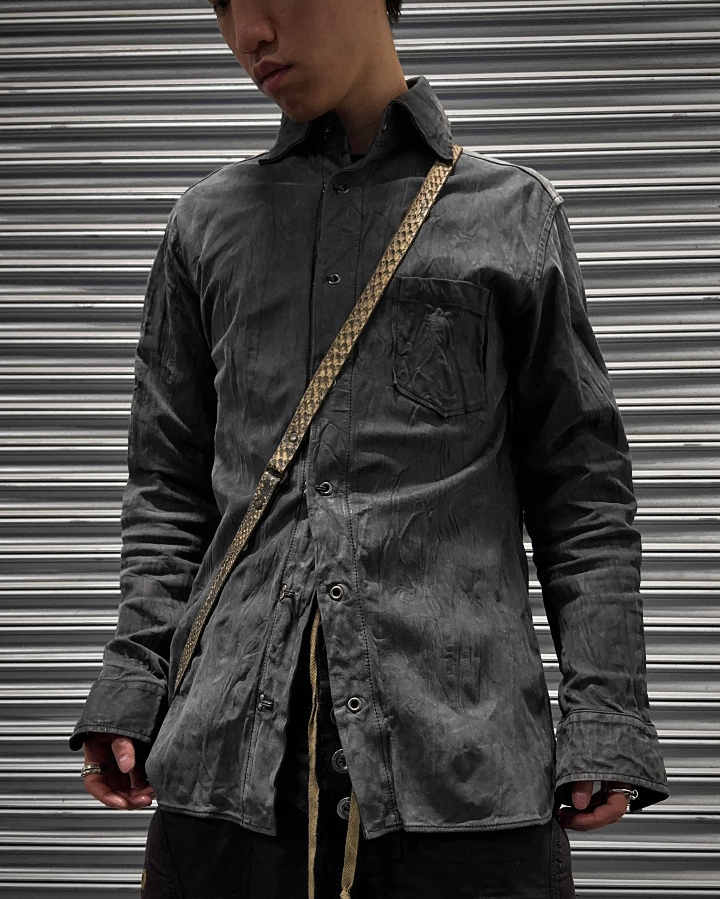 A person in a faded black Kyoji Maruyama Early 00s Washable Wrinkle Nubuck Leather Shirt with a front pocket and tan crossbody strap stands before a metallic, striped backdrop, their face partially visible, channeling Japanese minimalism.