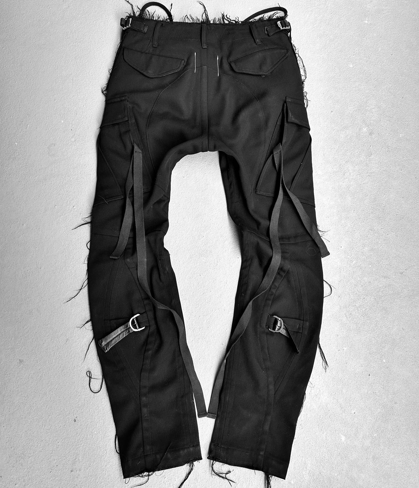 The TakahiroMiyashita TheSoloist. AW15 Calf-trimmed Wool ‘Bondage’ Straps Cargo Pants, influenced by avant-garde fashion, feature frayed edges and a distressed waist. Displayed on a gray surface with multiple straps, cargo pockets, and cuff buckles for an edgy appearance.