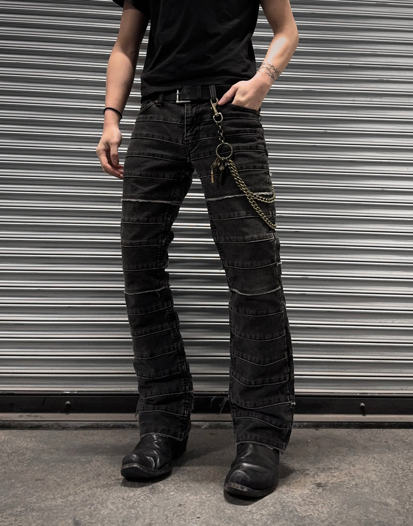 A person embodies punk rock style in Hysteric Glamours Early 00s Kinky Grey Flare Hagi Denim, with a chain-adorned black belt and boots. A bold tattoo graces their raised left arm as they pose confidently before a metal garage door, exuding effortless edginess.