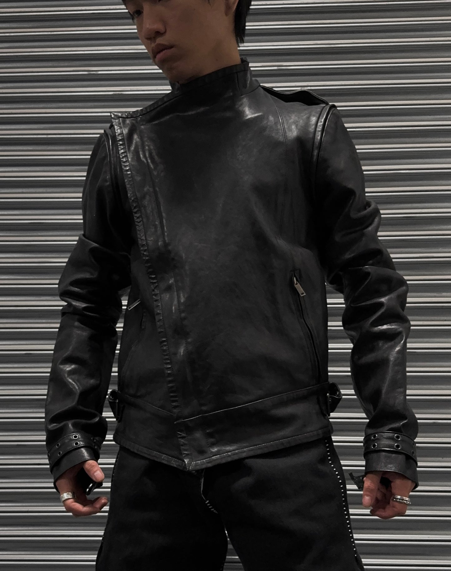Wearing the Gareth Pugh AW09 Detachable Sleeves Horse Leather Riders Jacket and black pants, a person stands before a metallic shutter, evoking avant-garde style while looking down and to the side with hands slightly raised.