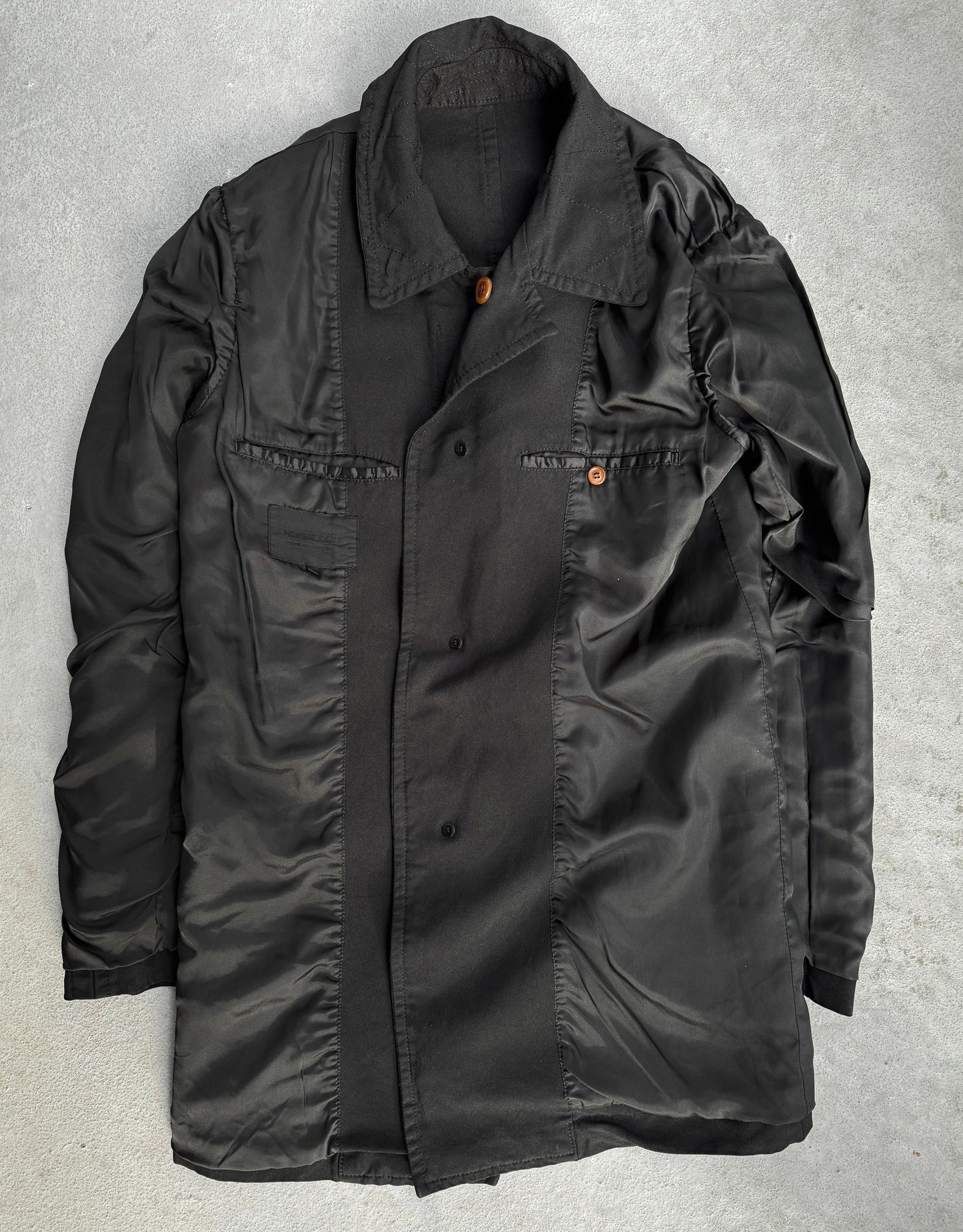 A black COMME des GARÇONS HOMME DEUX Early 00s Reversible Button-up Coat, showcasing a collared neck and two buttoned pockets, features an avant-garde blend of matte and glossy textures on a concrete background.