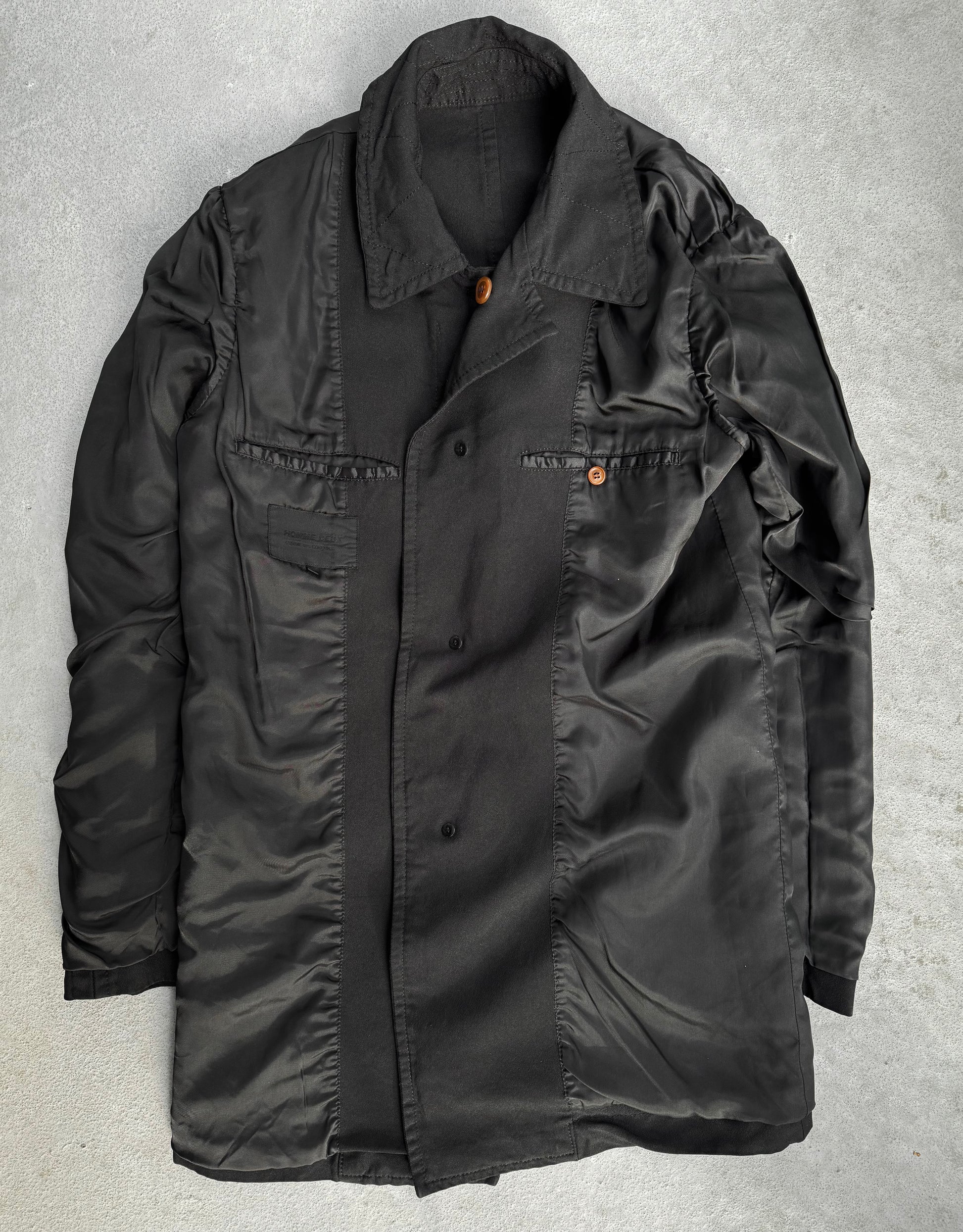 A black COMME des GARÇONS HOMME DEUX Early 00s Reversible Button-up Coat, showcasing a collared neck and two buttoned pockets, features an avant-garde blend of matte and glossy textures on a concrete background.
