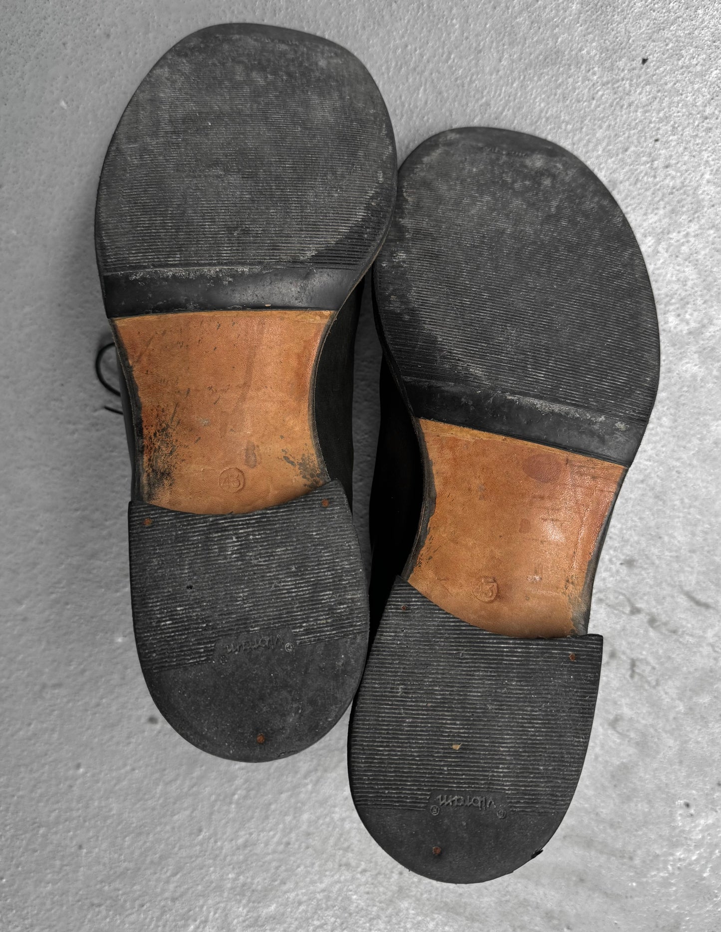 Pair of worn shoes with black soles and brown leather uppers on a gray concrete surface.