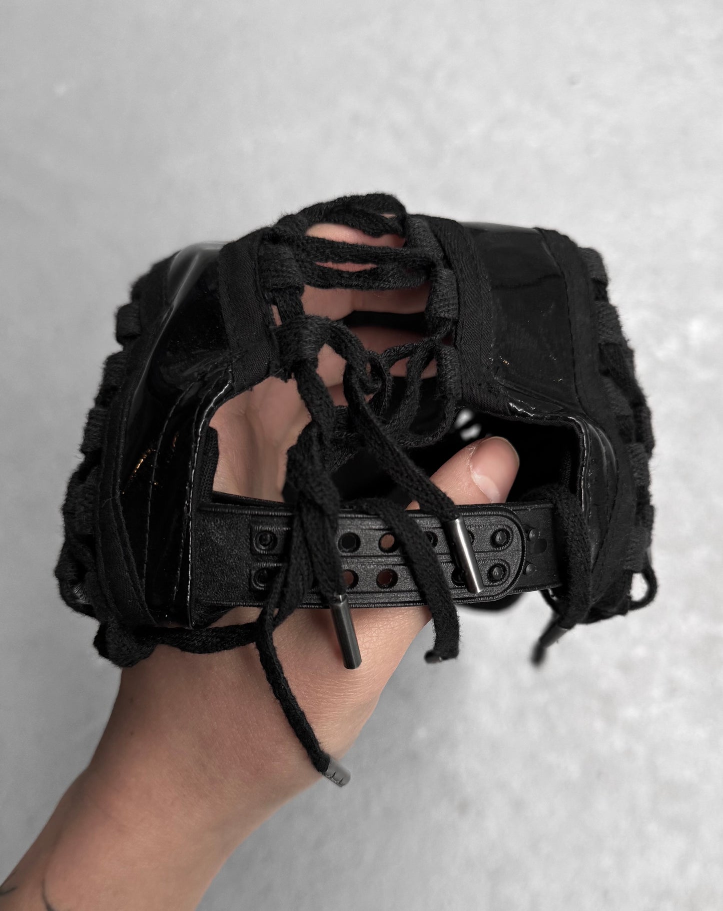 A hand holds a black KTZ sandal from Kokon to Zai, a London brand known for its East meets West style. The shoe showcases a woven design with lace details, rugged sole, and buckled strap against a plain grey backdrop.