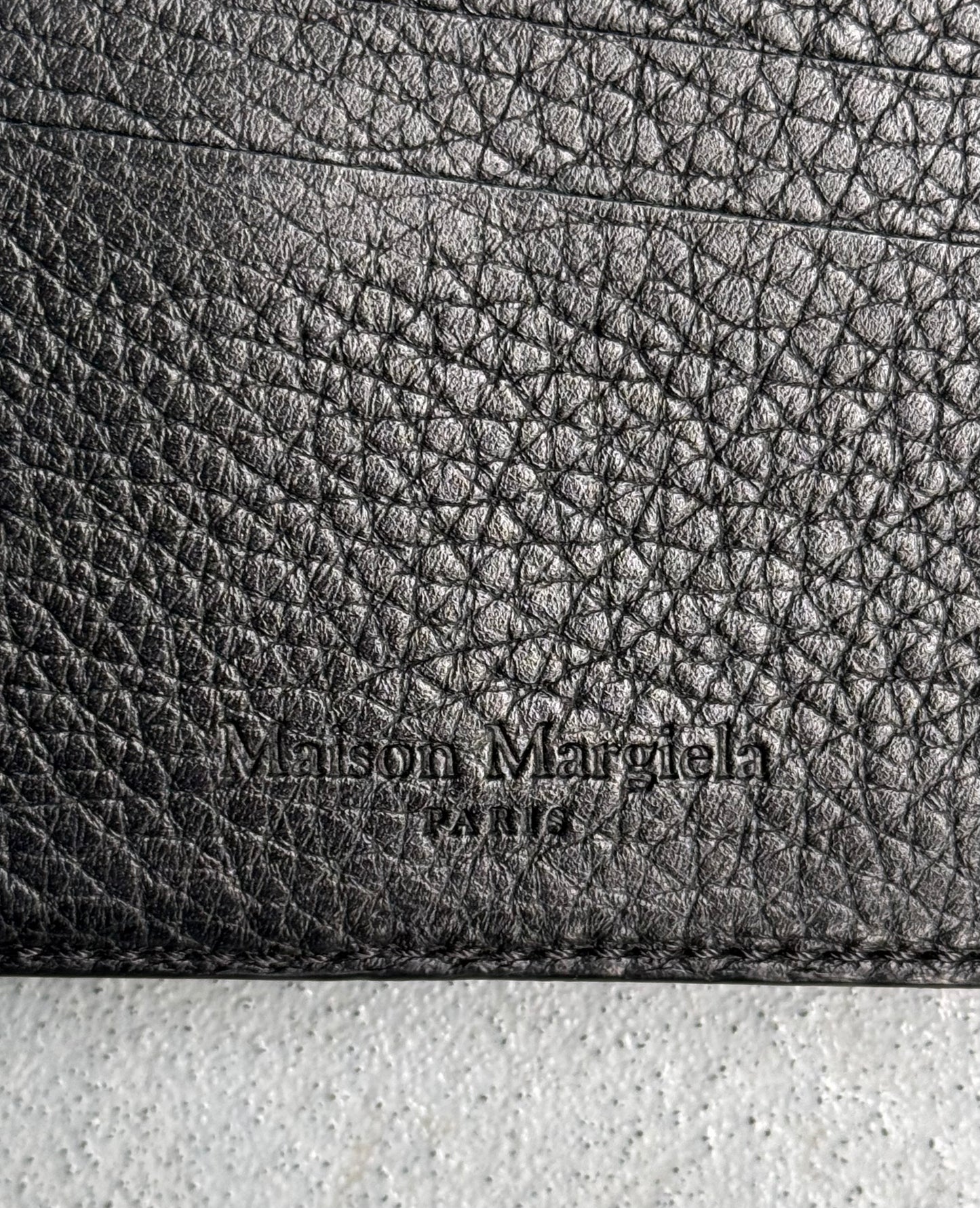 Close-up of black tumbled leather on the Maison Martin Margiela SS14 Bifold Wallet, featuring embossed Maison Margiela PARIS text. The grain and stitching reflect the brands avant-garde fashion legacy.