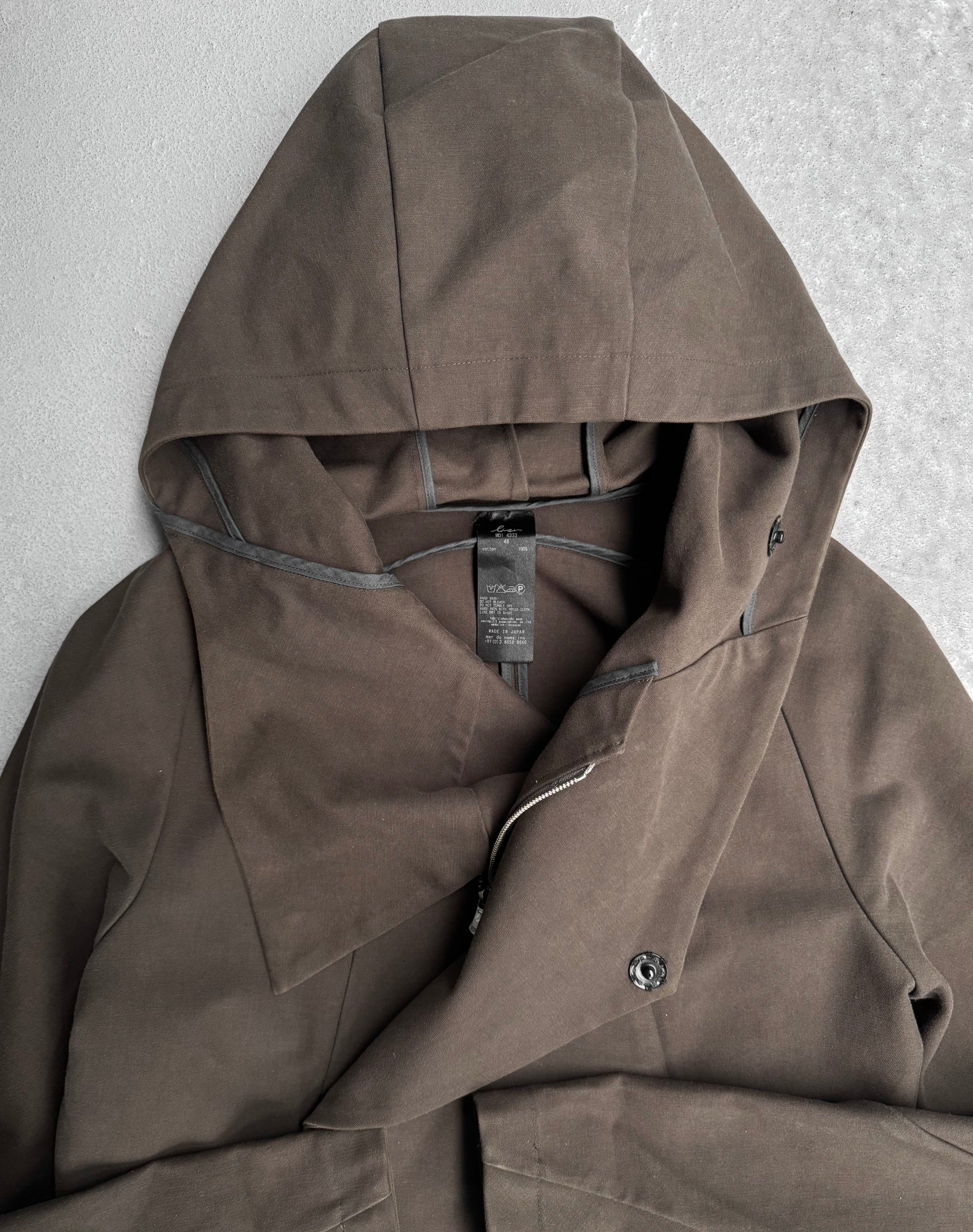 The lien AW12 Moleskin Asymmetrical Hooded Mod Coat in dark brown, with a unique Japanese-inspired design and asymmetrical button closure, is displayed flat. The spread hood shows a black brand label against a light gray background, highlighting its modern elegance.