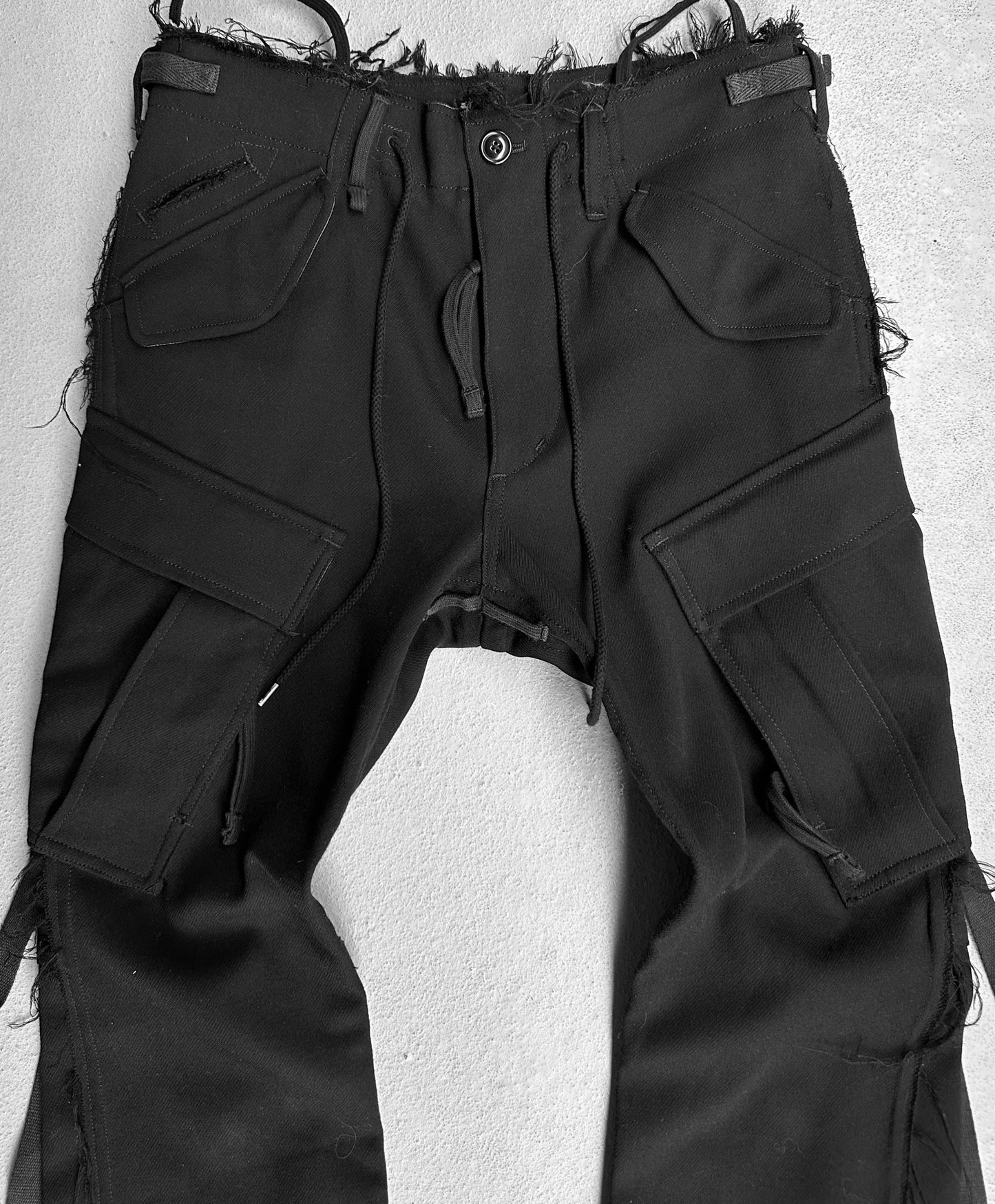 TakahiroMiyashita TheSoloist. AW15 Calf-trimmed Wool Bondage Straps Cargo Pants, with frayed edges and multiple pockets, echo Takahiro Miyashitas avant-garde style. Featuring a distressed waist, zipper details, and belt loops, they complete the edgy look on a light surface.