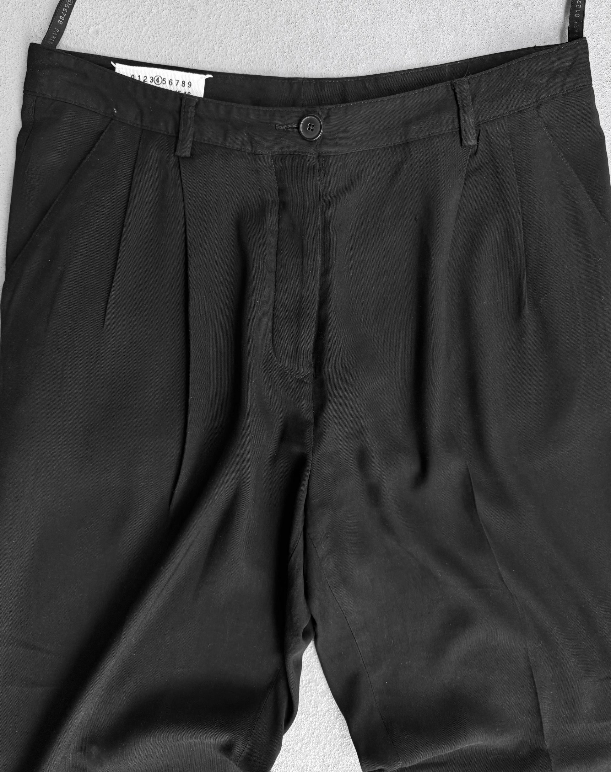 Maison Martin Margiela Early 00s Double Pleated Dress Pants, showcasing avant-garde fashion, are displayed on a light gray background. They feature a single button and zipper, visible belt loops at the waistband, and a subtly visible size tag that hints at anti-branding simplicity.