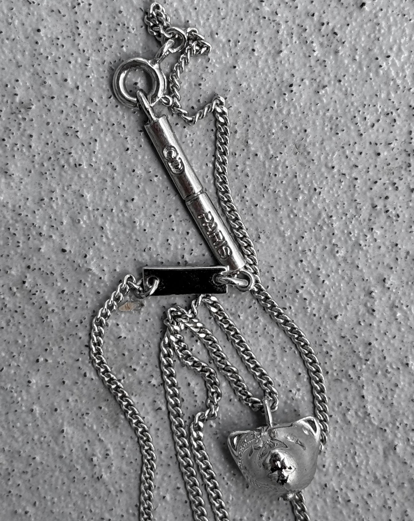 The DIOR HOMME Early 00s Valentine’s Day Necklace features a silver teddy bear pendant and an engraved bar, reminiscent of Hedi Slimanes era, with delicate links artfully tangled on a speckled gray surface.
