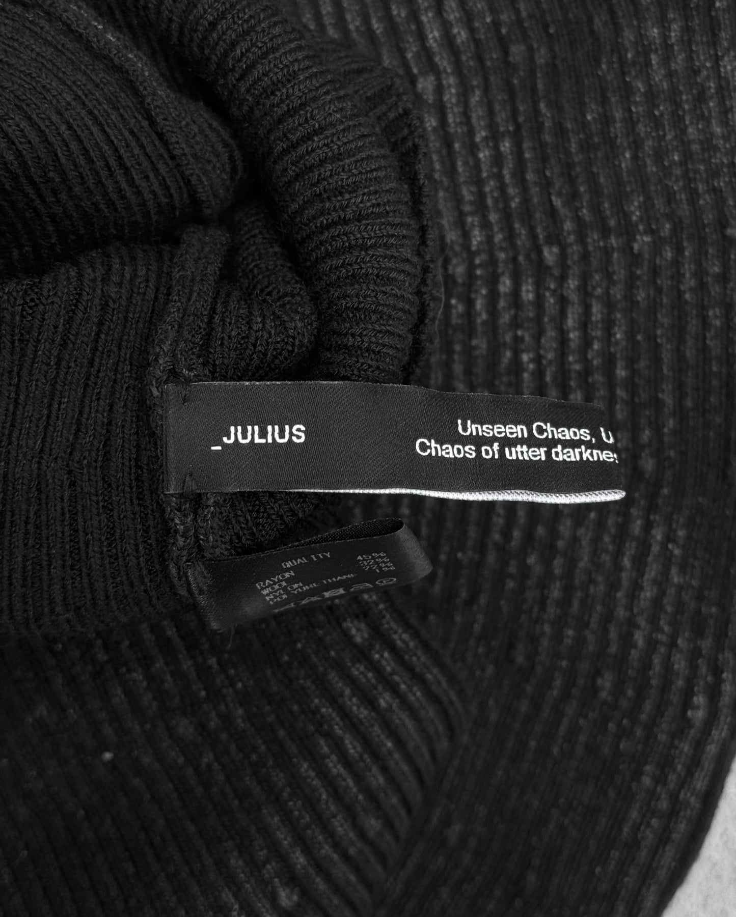 A close-up of DRIEW’s Julius_7 AW24-25 “rift;” Curved Wax Coated Gloves in flawless black ribbed fabric, featuring a “_JULIUS” label with partially visible text: “Unseen Chaos… utter darknes…”. One size fits most.