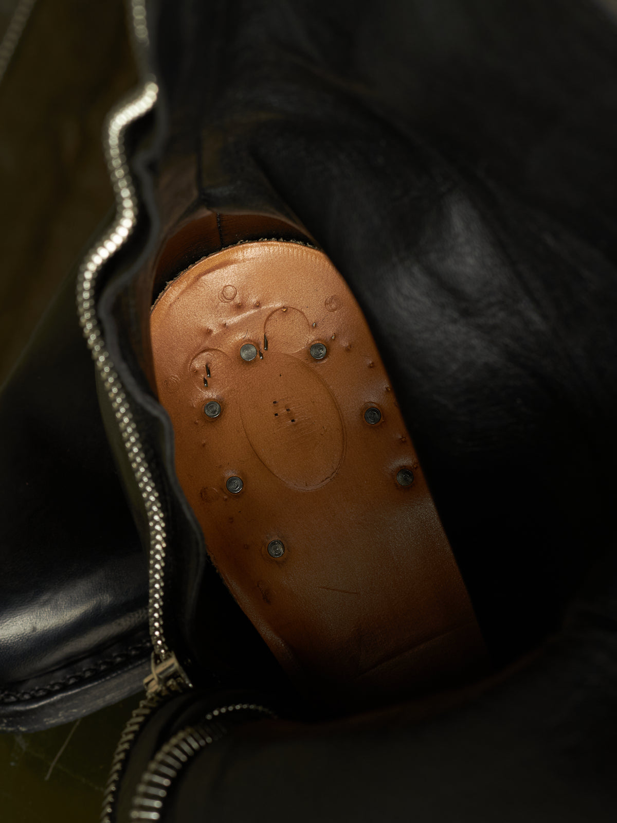 Close-up of a black leather boot or bag opening, showing a tan leather insole with metal studs and a circular embossed detail.