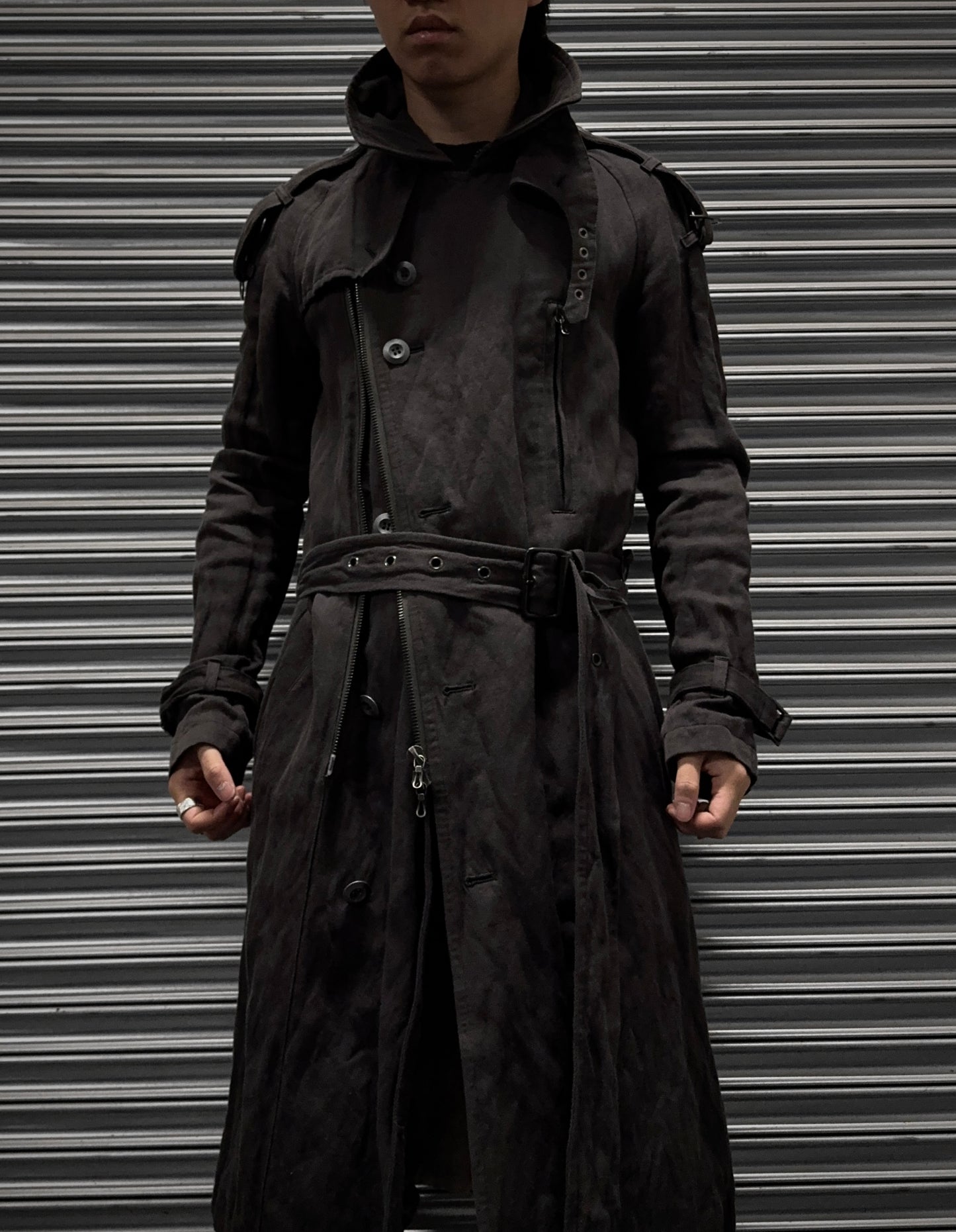 A person in an ATTACHMENT by Kazuyuki Kumagai Raglan Doctor Trench Coat stands before a closed metal shutter, their face partly shadowed and hands resting at their sides.