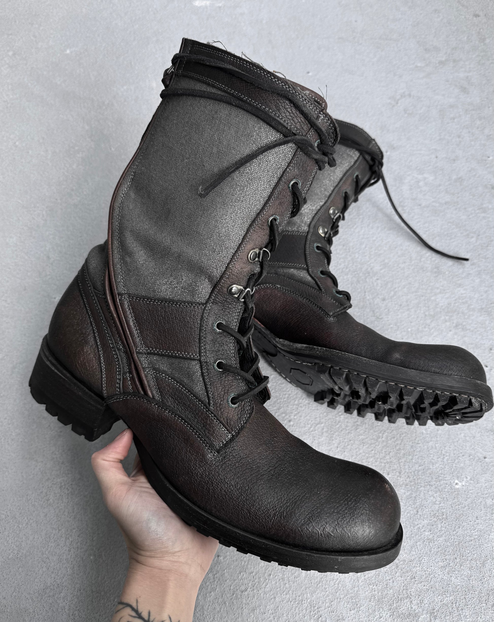 A person with a wrist tattoo holds a pair of Kyoji Maruyama Early 00s Brown Tornado Zip Combat Boots, made from rugged sheepskin leather, featuring thick soles and lace-up fronts, against a neutral background.