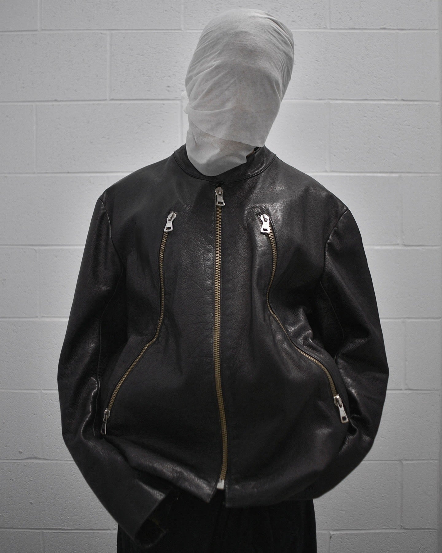 A person in front of a white brick wall wears the Maison Martin Margiela SS00 Black Cowhide Leather 5-zip Rider Jacket by Maison Martin Margiela, their face fully wrapped in white cloth—a bold statement in avant-garde fashion.