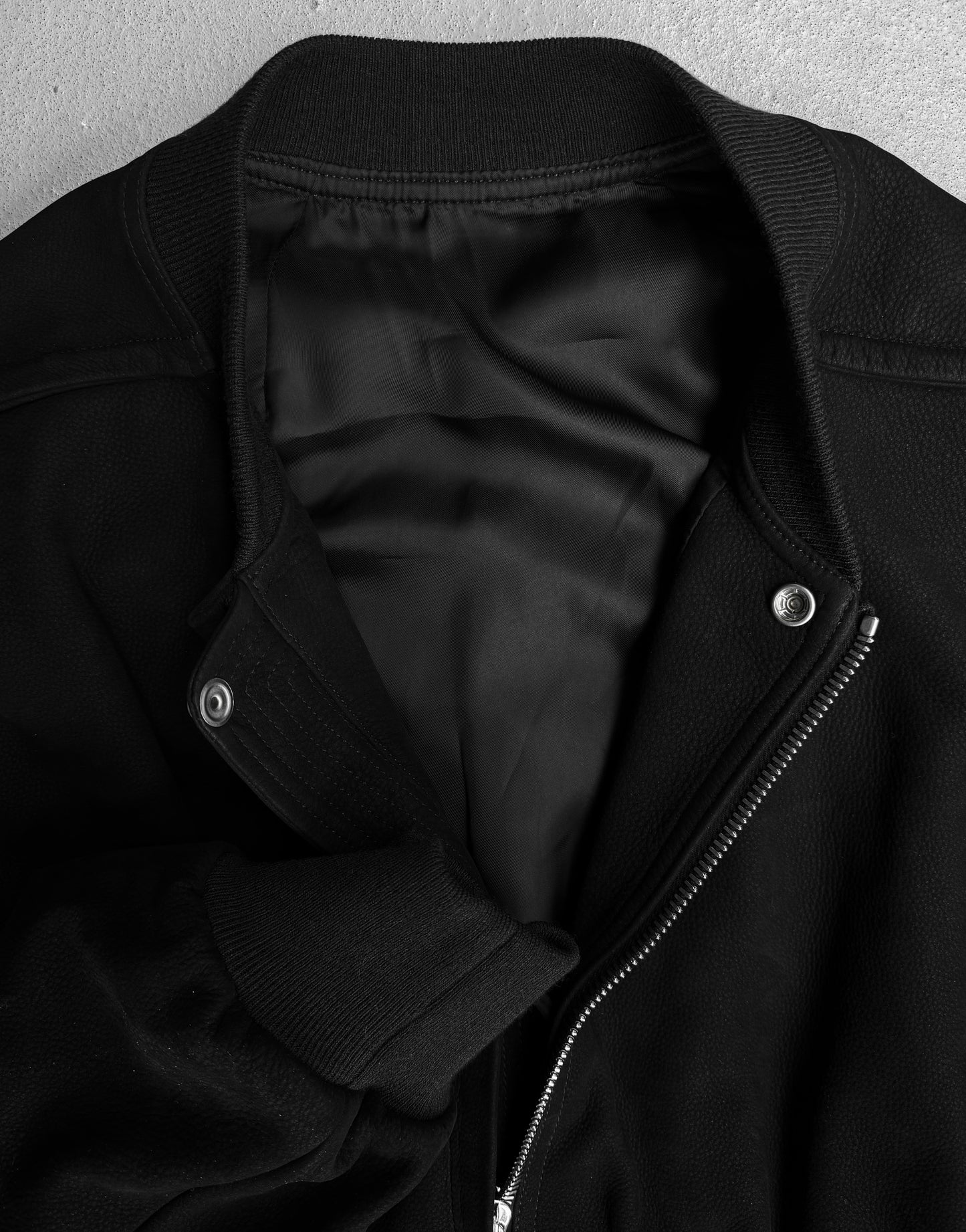 A close-up of the Rick Owens SS24 “LIDO” Nubuck Leather Cropped Flight Bomber Jacket features a zipper, snap buttons, ribbed collar, cuffs, and hems. Partially unzipped, it reveals a smooth black lining in true avant-garde Rick Owens style.