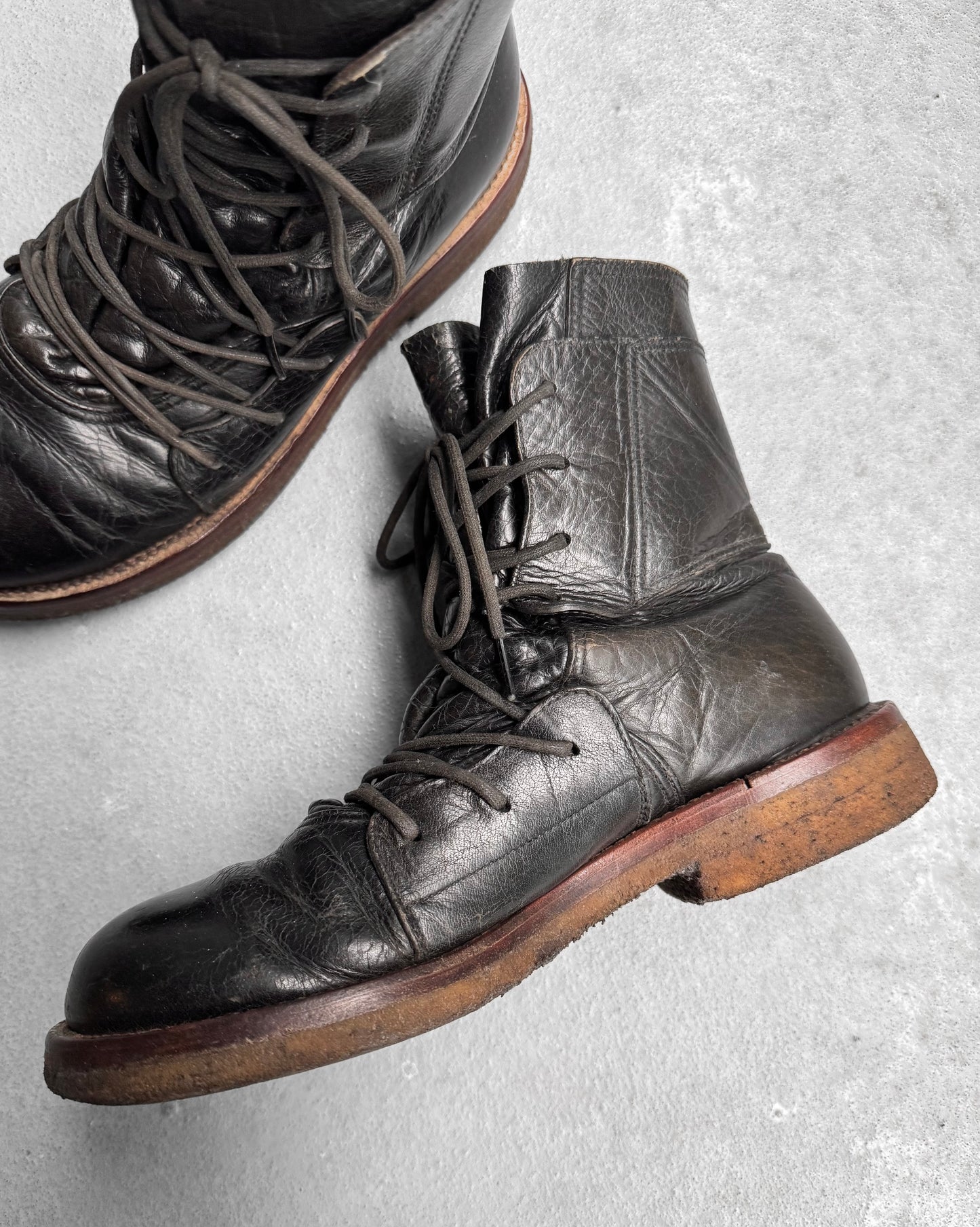 The MiharaYasuhiro Early 00s Multi-laced Crepe Sole Boots by Mihara Yasuhiro, with black tops, brown soles, and long laces, are set against a gray textured background. The boots exhibit rugged deconstructed elements of avant-garde fashion with a slightly scuffed appearance, indicating frequent use.
