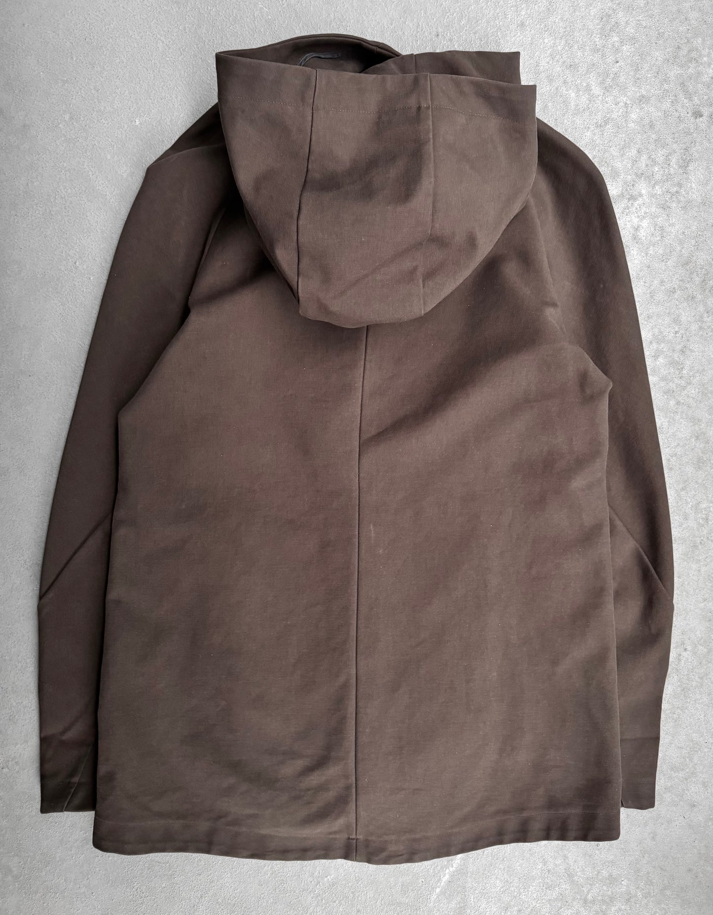 The lien AW12 Moleskin Asymmetrical Hooded Mod Coat in brown is displayed back-first on a textured gray surface. It features an elegant centered seam and long sleeves, merging traditional Japanese fashion with contemporary flair.