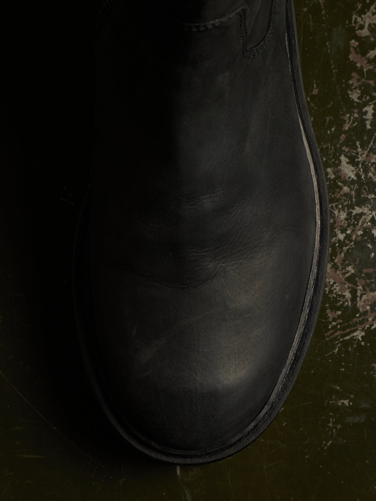 A close-up, top-down view of a black leather boot on a dark, textured green-brown surface.