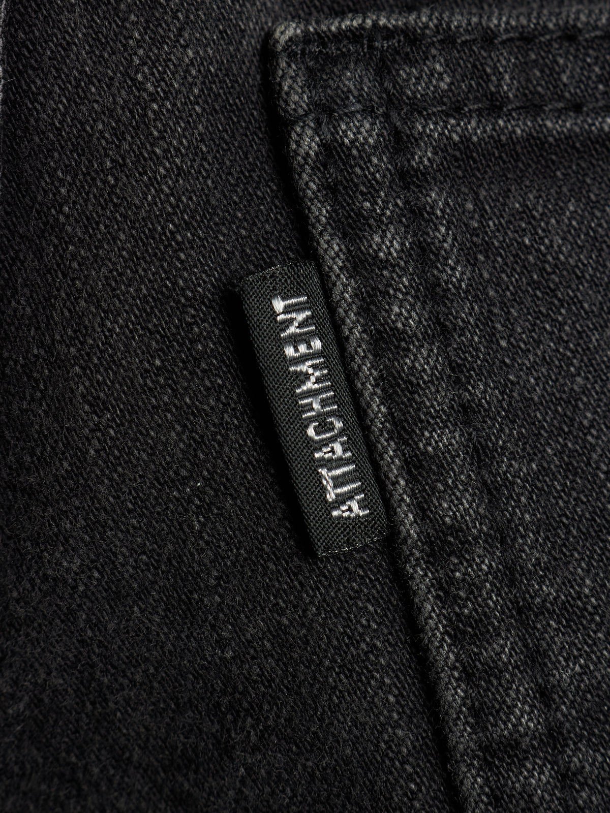 Close-up of black denim fabric with a small black brand tab reading ATTACHMENT stitched on the side.