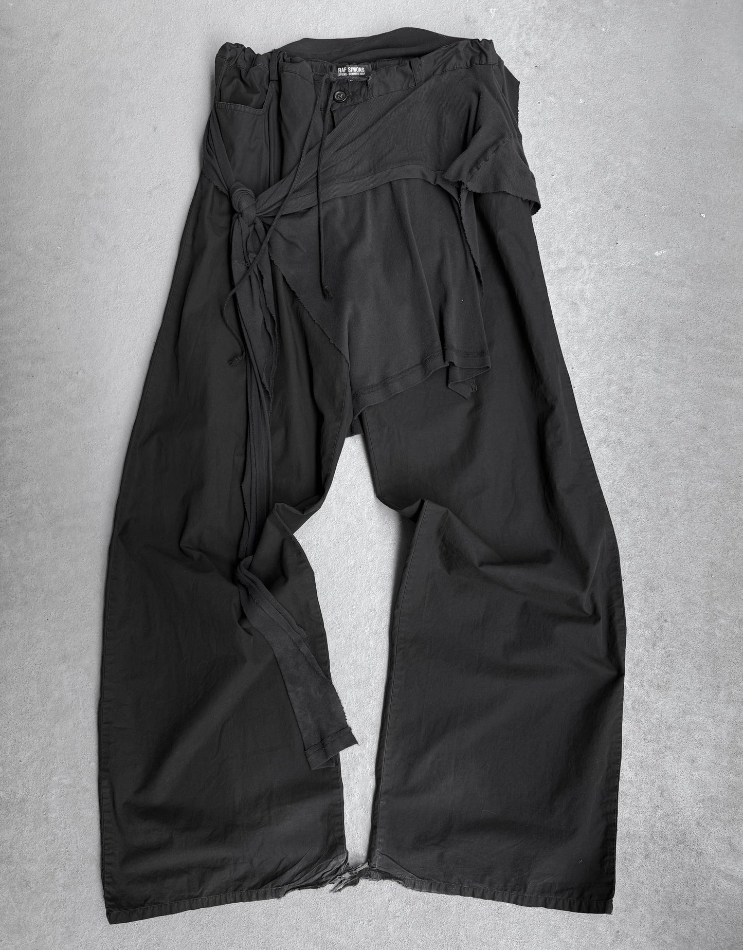 RAF SIMONS SS04 “May the Circle Be Unbroken” Reconstructed ‘Zombie’ Wide Leg Pants, in black with wrap detail and loose fabric ties, feature a relaxed fit and slightly crinkled texture for minimalist style. Laid flat on a light gray surface.