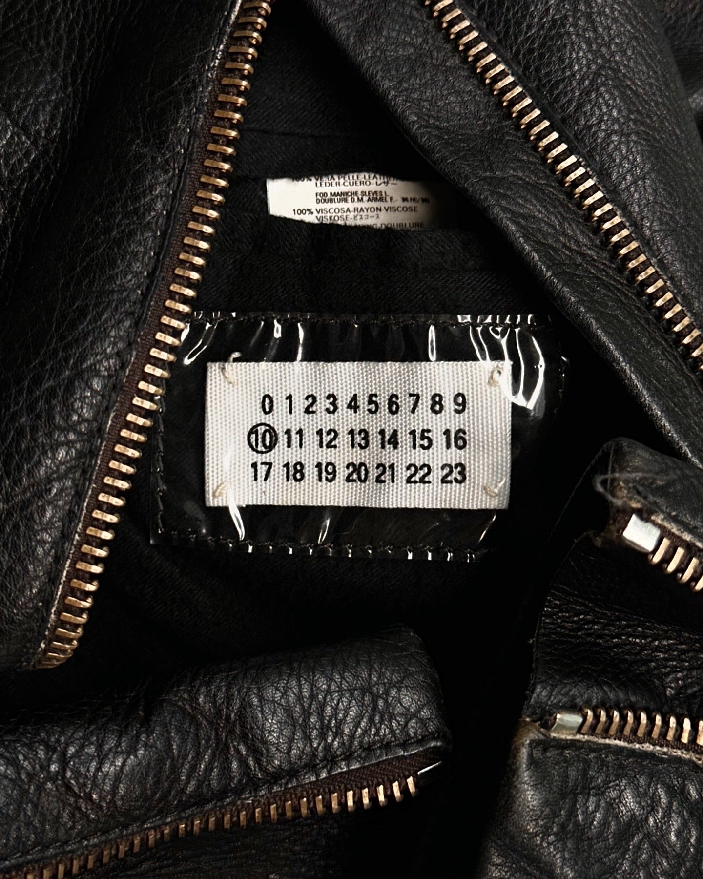 A close-up of the Maison Martin Margiela SS00 Black Cowhide Leather 5-zip Rider Jacket shows silver zippers and an inner label featuring numbers 0–23 with 10 circled, a signature detail of Maison Martin Margiela.