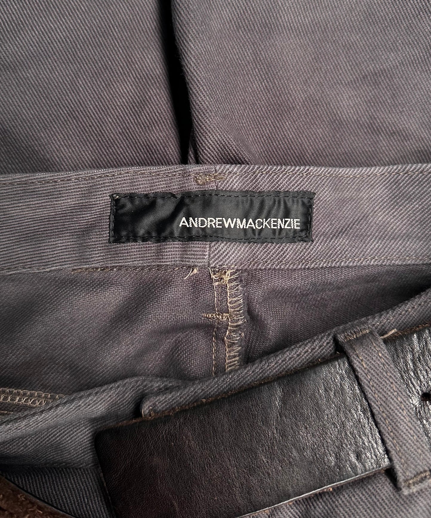 Close-up of ANDREW MACKENZIE Early 00s Stud Darted Knee Denim, showing grey fabric with a black ANDREW MACKENZIE label on the waistband, folded knee detailing, and a partially visible brown leather belt.