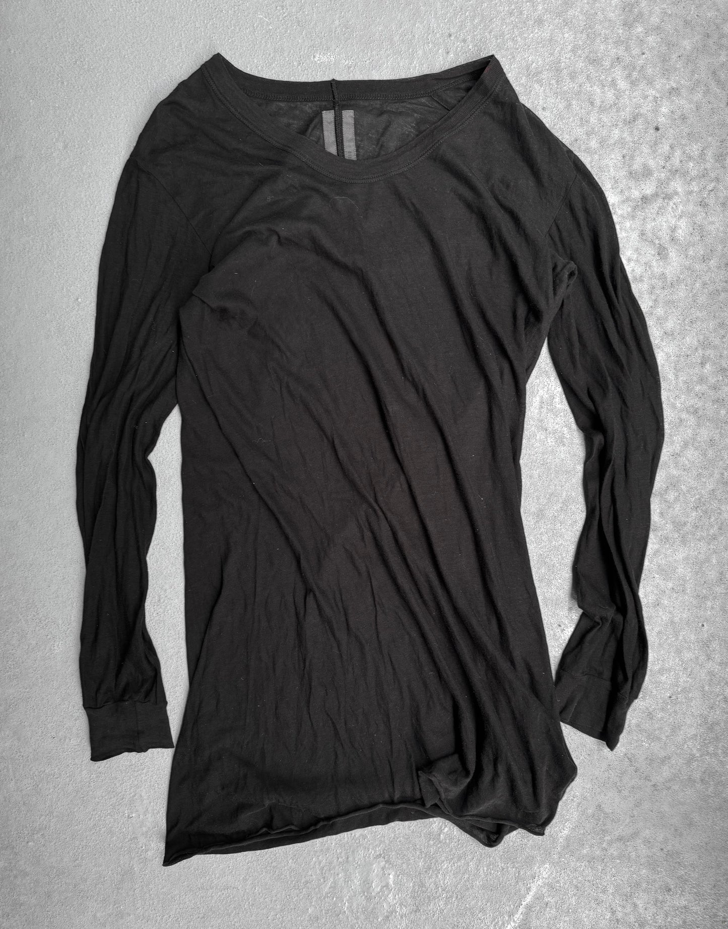 The Rick Owens SS10 “RELEASE” Black Sheer Long-sleeves lies flat on a light gray textured surface, showing visible wrinkles and a slightly bent sleeve, reflecting the brands signature contemporary style.