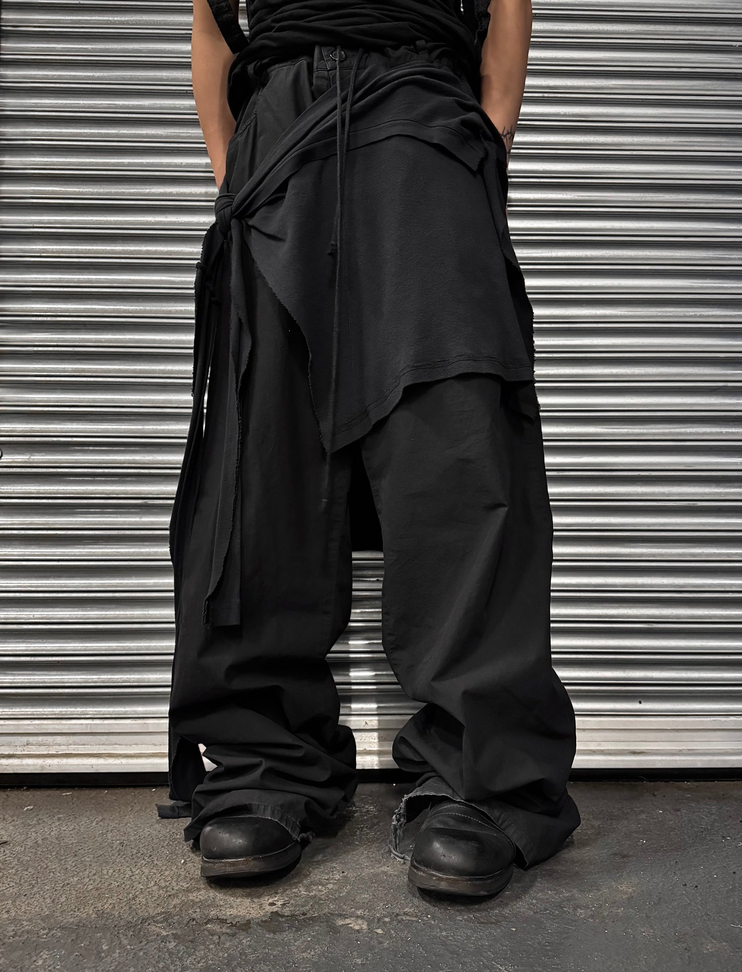 A person stands before a metal shutter, channeling poetic minimalism in loose, layered RAF SIMONS SS04 May the Circle Be Unbroken Reconstructed Zombie Wide Leg Pants and shoes—a subtle homage to RAF SIMONS’ avant-garde style.