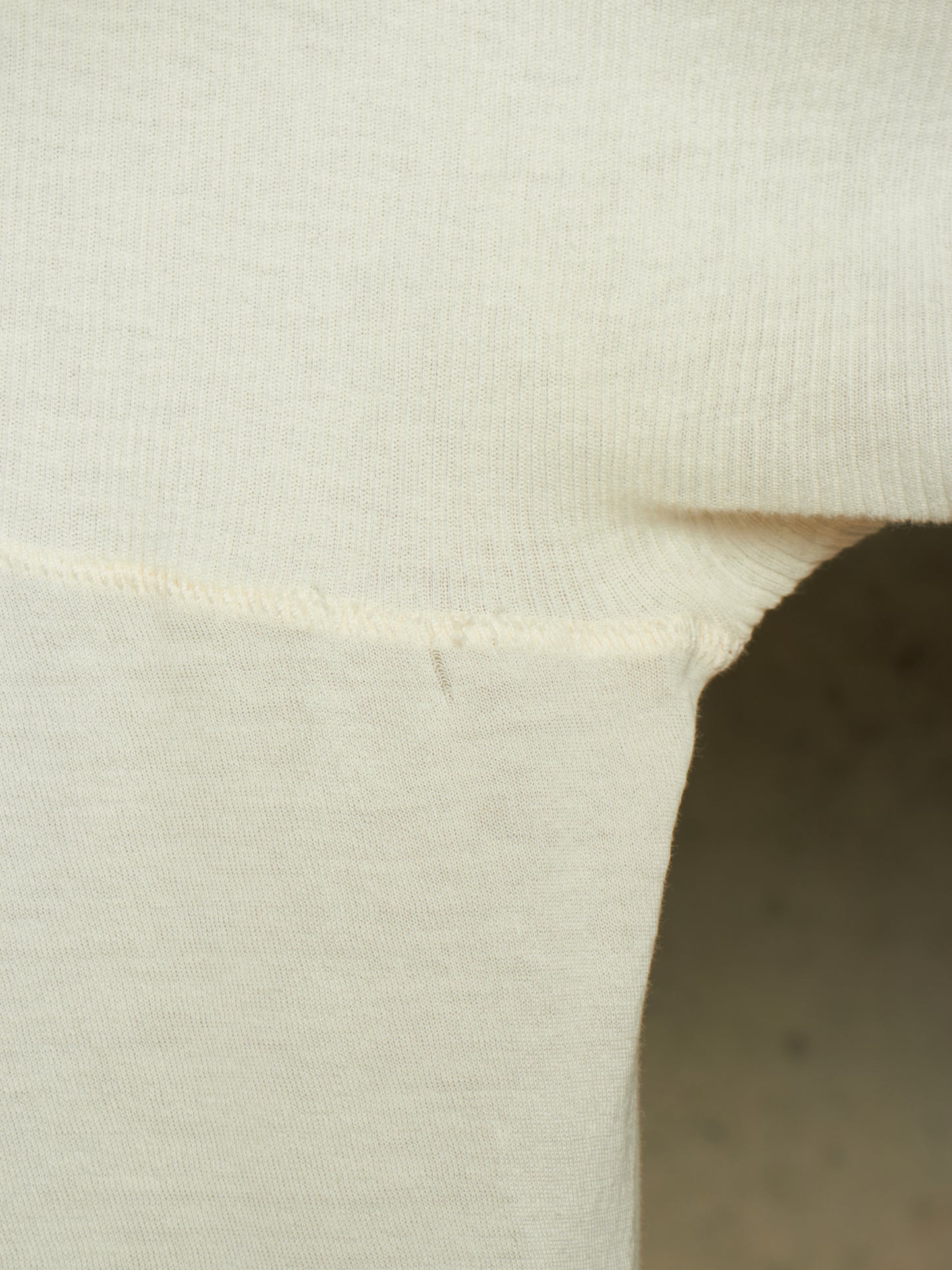 Close-up detail of a cream-colored knit garment, showing the ribbed texture and a folded seam against a neutral background.