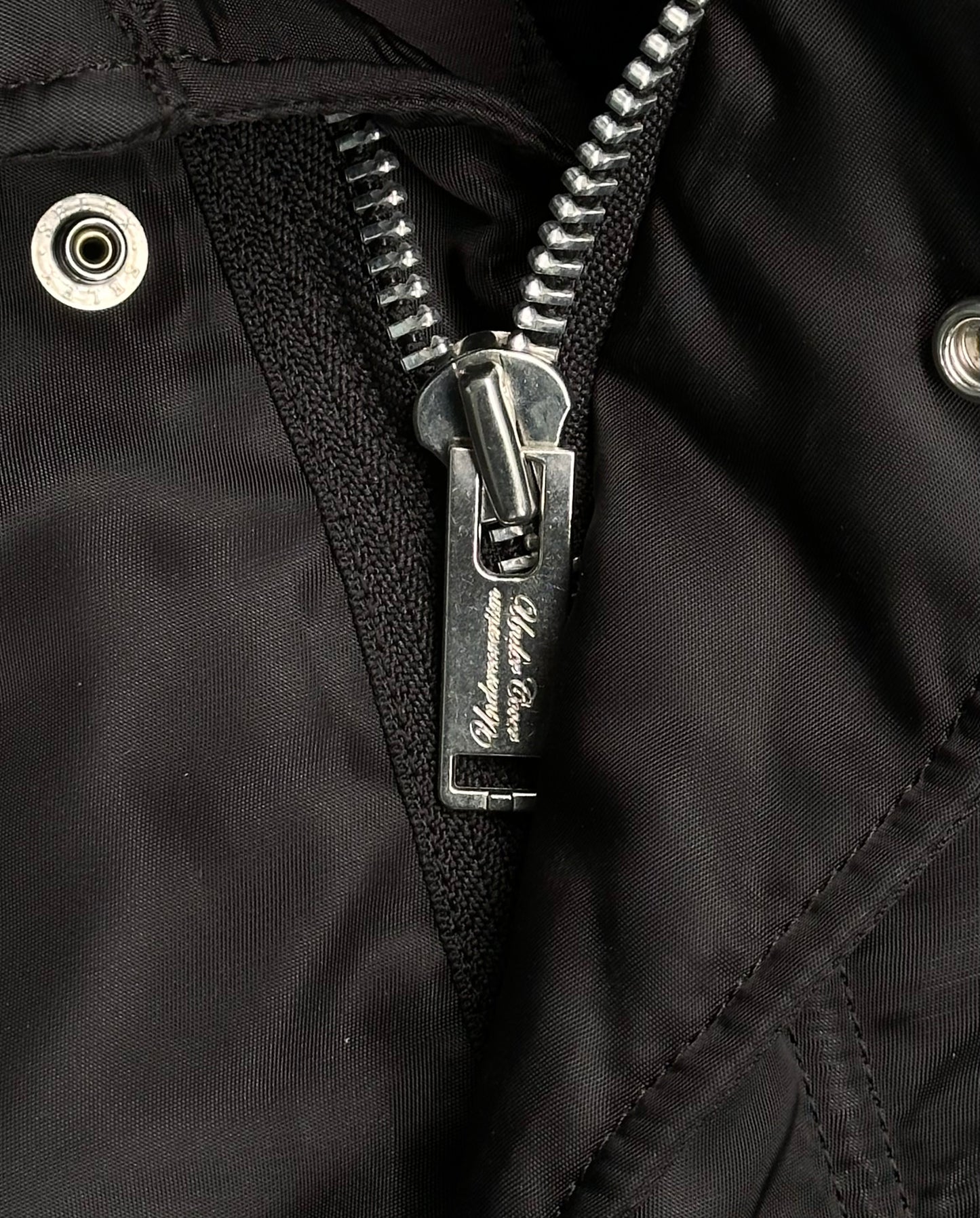 Close-up of the UNDERCOVER AW16 ‘Borremans’ Full-zip Fishtail Nylon Parka, showing its black fabric, large metallic silver zipper with engraved text, and two silver grommets—a signature detail from designer Jun Takahashi.