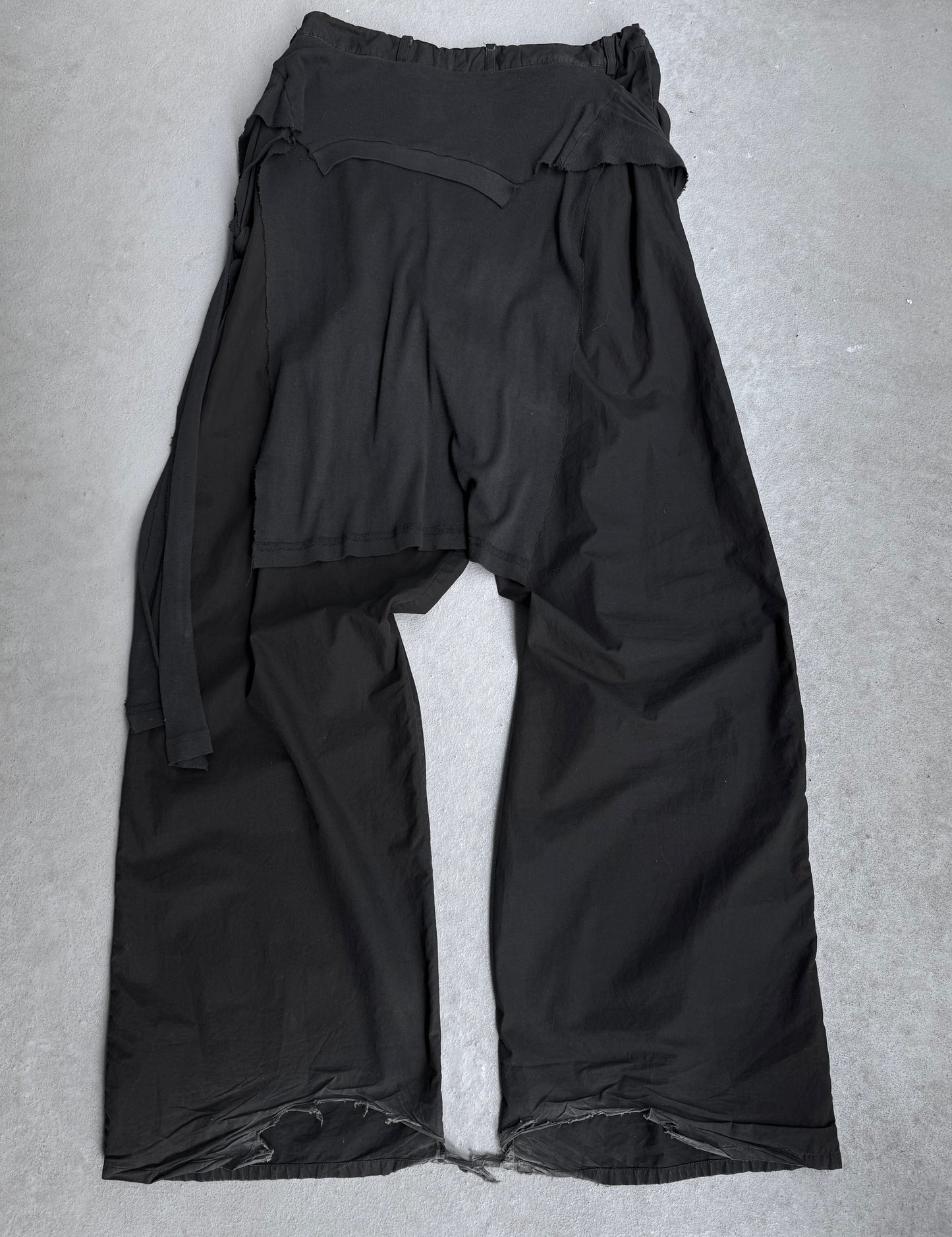 RAF SIMONS SS04 “May the Circle Be Unbroken” Reconstructed ‘Zombie’ Wide Leg Pants feature loose black construction, raw hem edges, an uneven waistband, and a long fabric belt—showcasing poetic minimalism on a gray concrete surface.