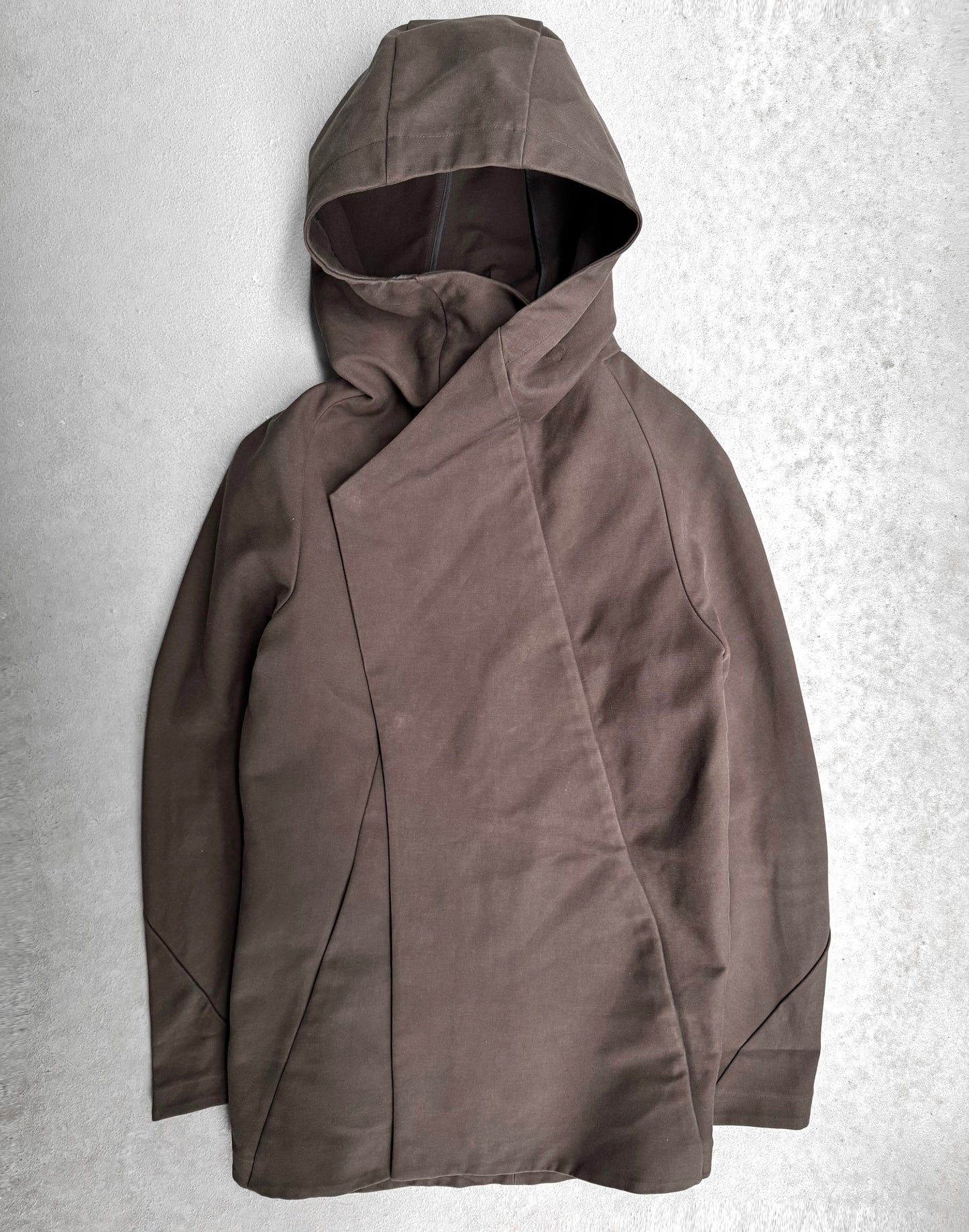 The lien AW12 Moleskin Asymmetrical Hooded Mod Coat, in brown, lies on a light gray surface. It has an avant-garde design with a diagonal closure, echoing Liens Japanese fashion with its soft and slightly textured fabric.