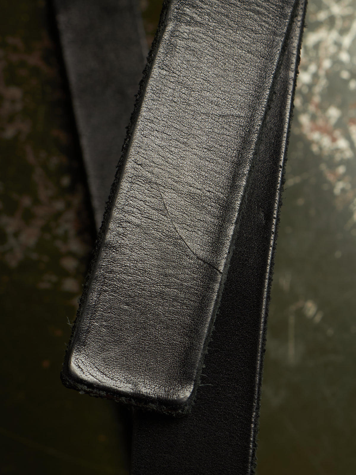 Close-up of a black leather strap, likely a belt or strap accessory, showing the textured grain on the top surface and stitched edges against a dark, mottled background.