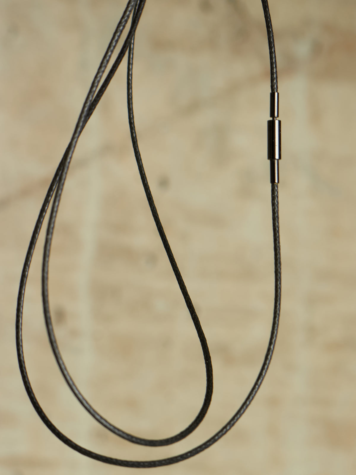 A close-up of a black braided cable necklace or cord accessory with a small metallic connector, shown against a warm beige textured background.