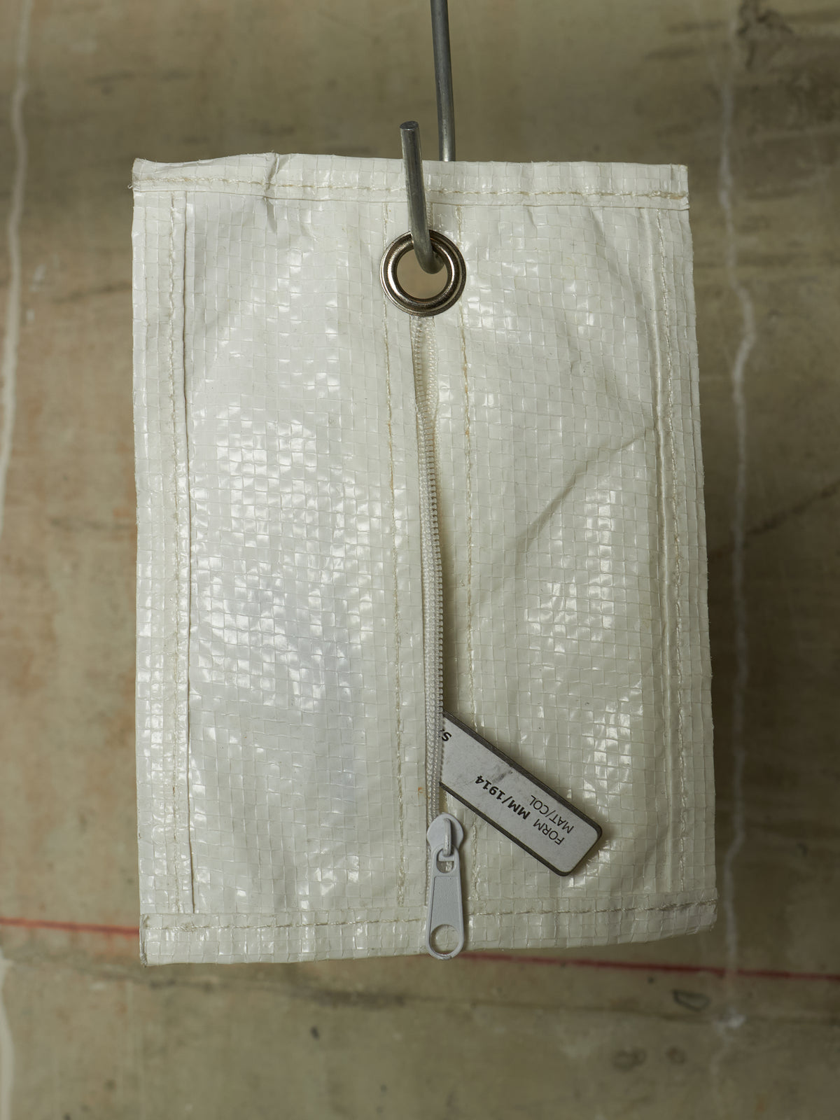 An off-white woven canvas utility pouch with a vertical zipper and a metal grommet at the top, hanging against a textured concrete wall.