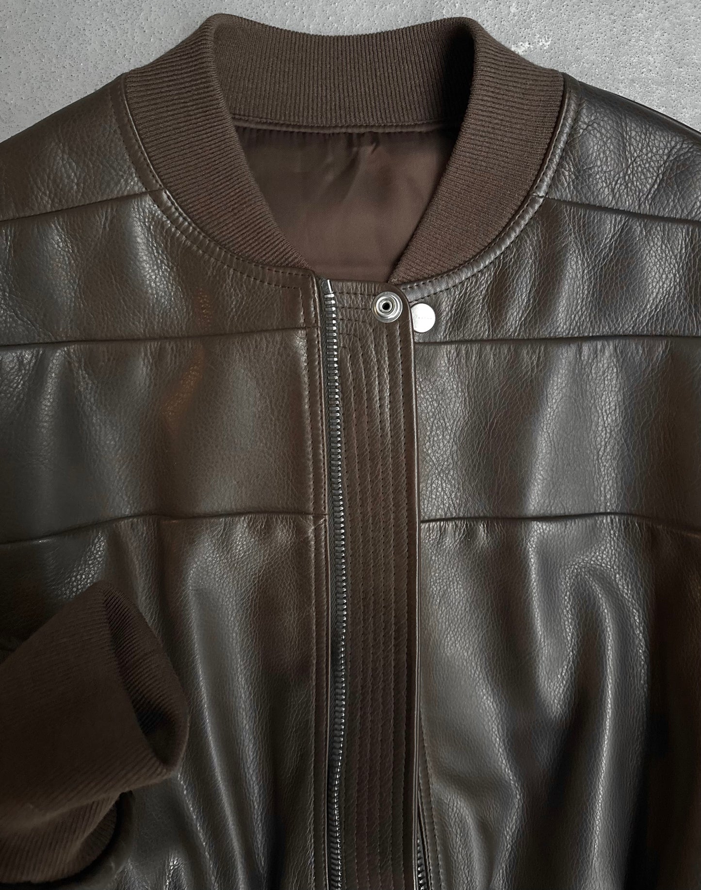 A close-up of the Rick Owens AW23 LUXOR Girdered Leather Bomber Jacket, crafted in dark brown calfskin with a ribbed knit collar and cuffs, snap button at the neck, and vertical stitching on the front.