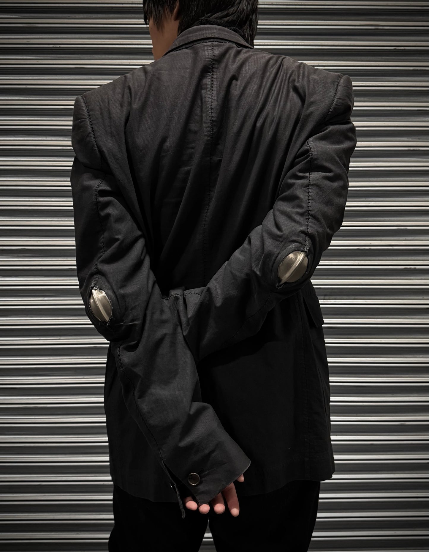 Person wearing a black jacket with metallic elbow pads against a metallic shutter background