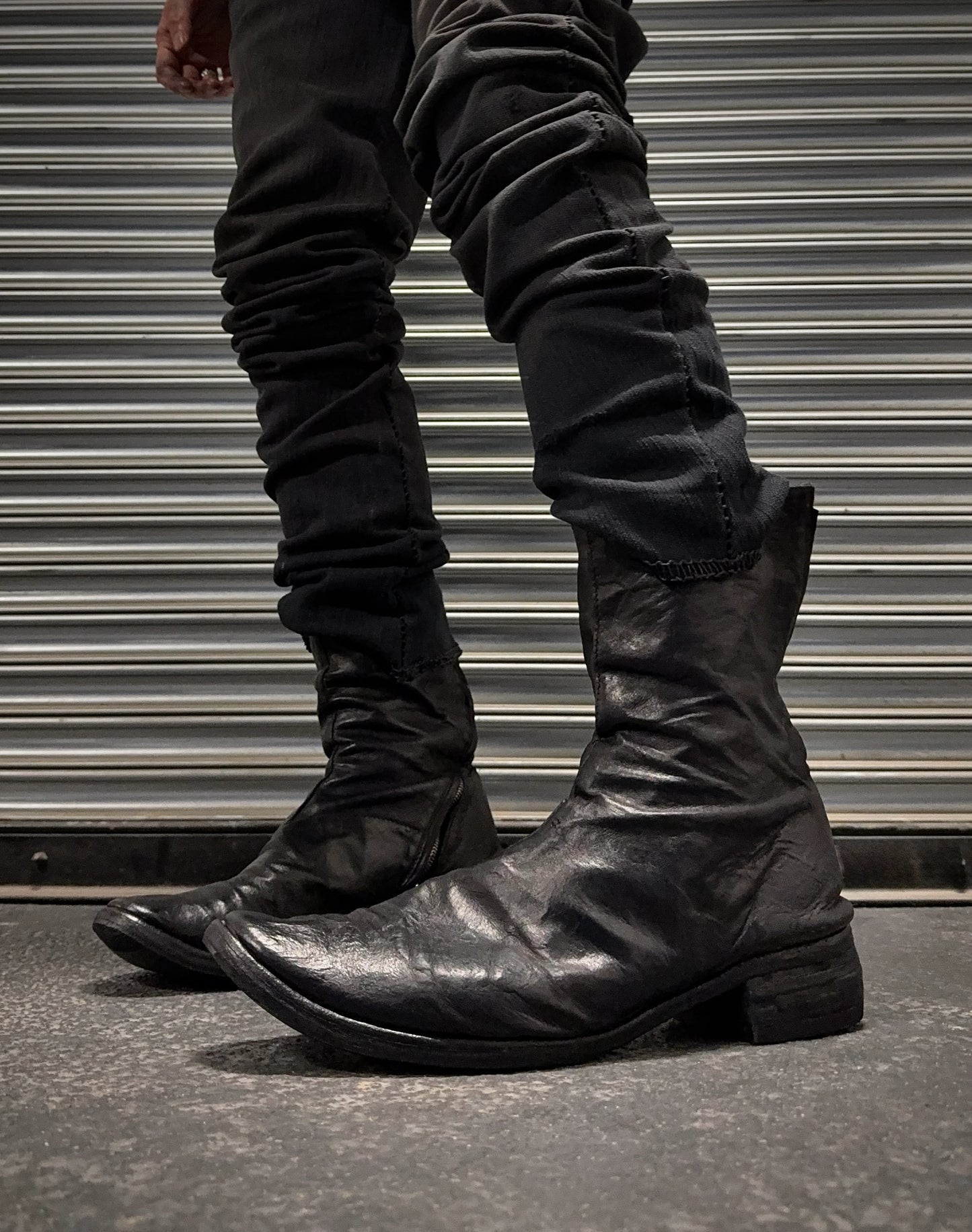 Black leather boots worn with dark pants against a metallic shutter background