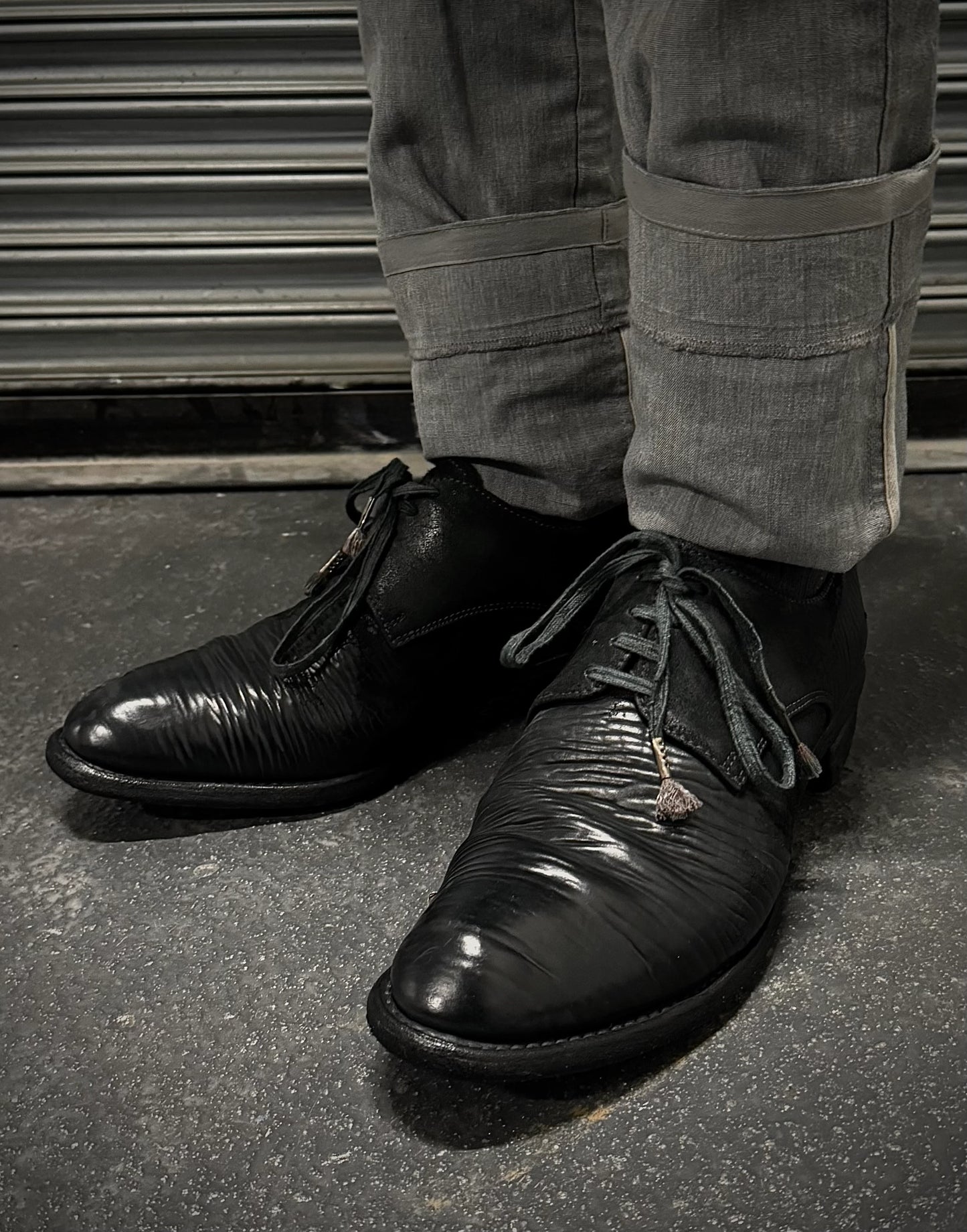 Black leather shoes worn with gray pants on a dark surface.